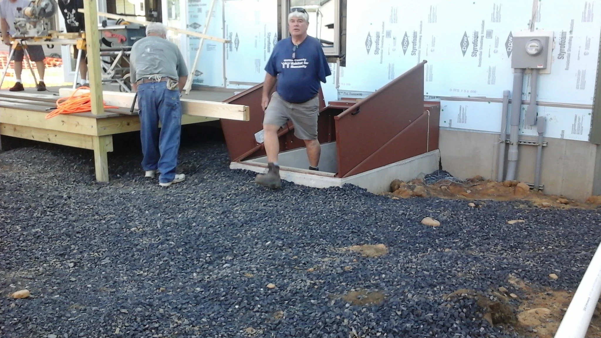 Construction workers are working on a building. One is stepping down into an underground area while another is working on a raised platform. Building materials and tools are present.