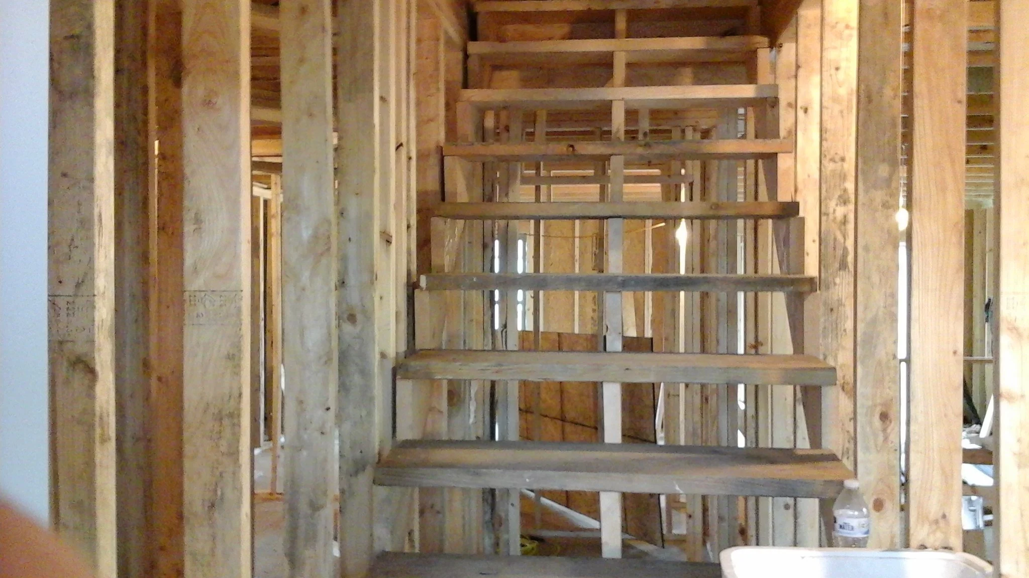 Construction site showing wooden framing and stairs in progress, with a water bottle and part of a sink in the foreground.