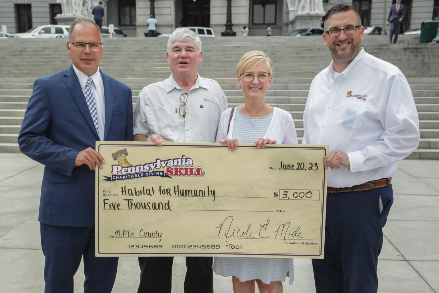 Four people standing in front of steps holding a large check for five thousand dollars made out to Habitat for Humanity in Mifflin County, with the date June 20, 2023. The group includes three men and one woman, all smiling.