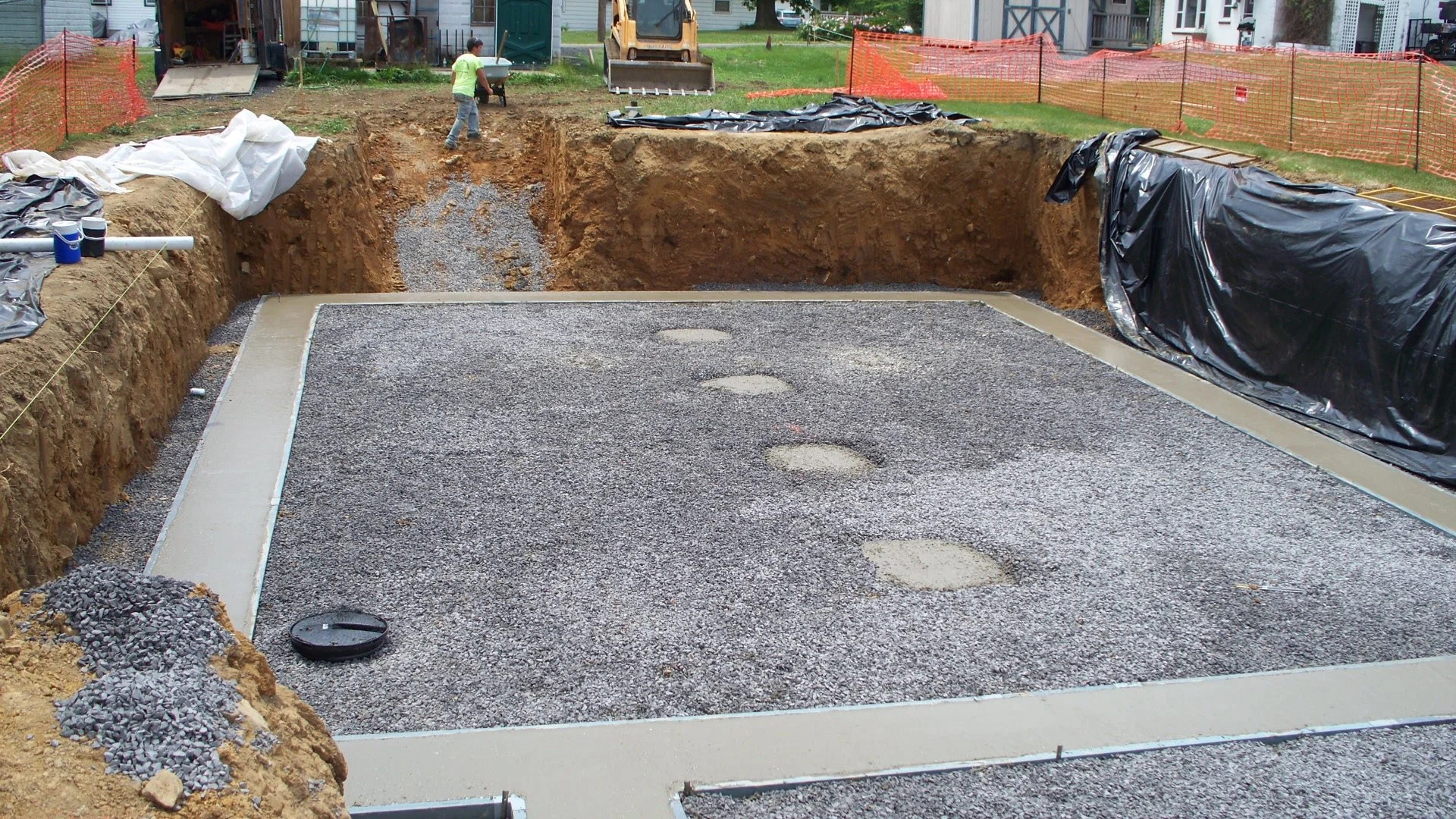View of an outdoor construction site where a foundation is being prepared, with gravel, concrete forms, and footprints in the gravel, surrounded by orange safety fencing.