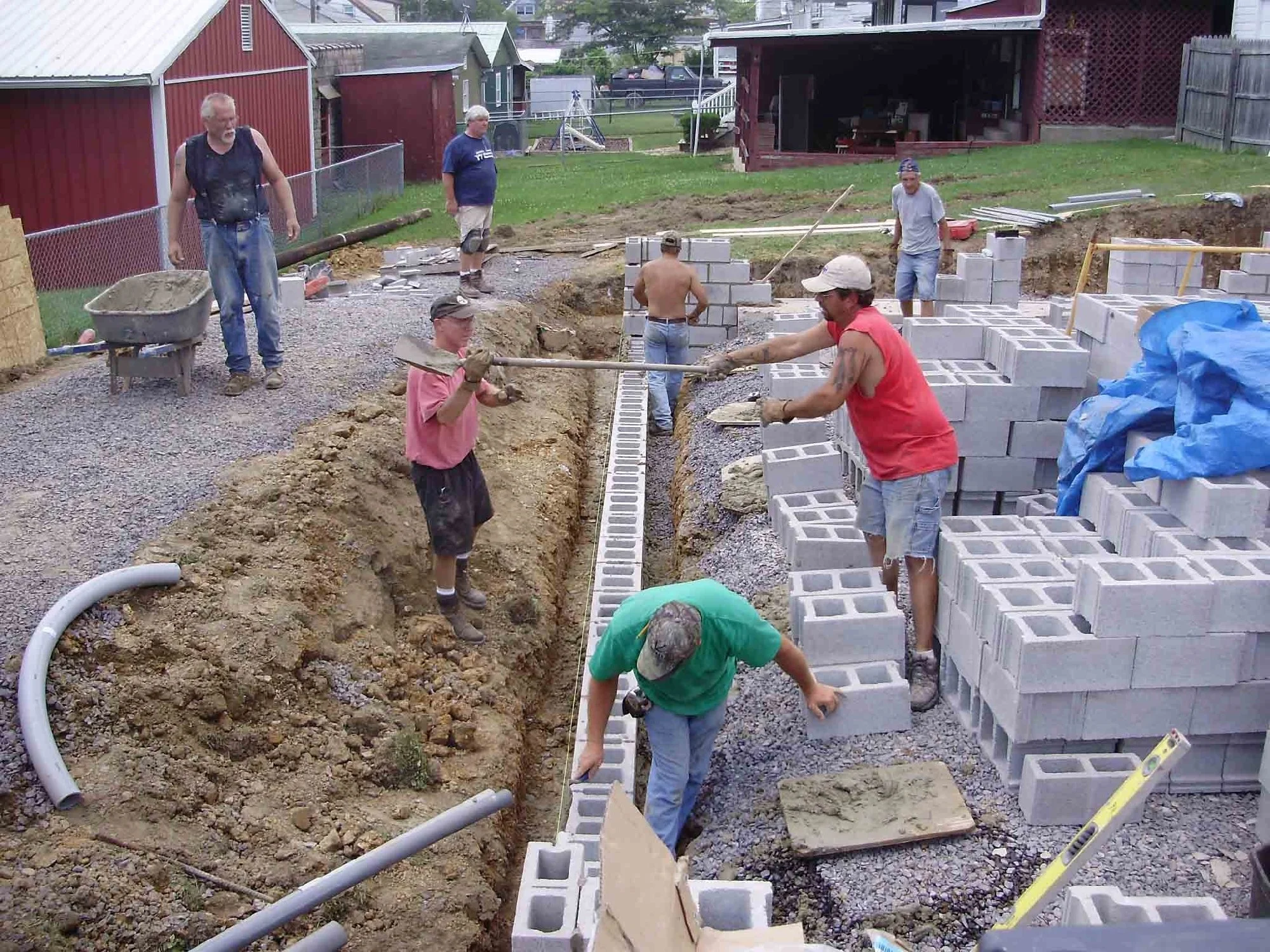 A group of construction workers building a brick wall outdoors. Some are working in a trench, while others are handling bricks and tools. The area is surrounded by a fence and residential buildings.