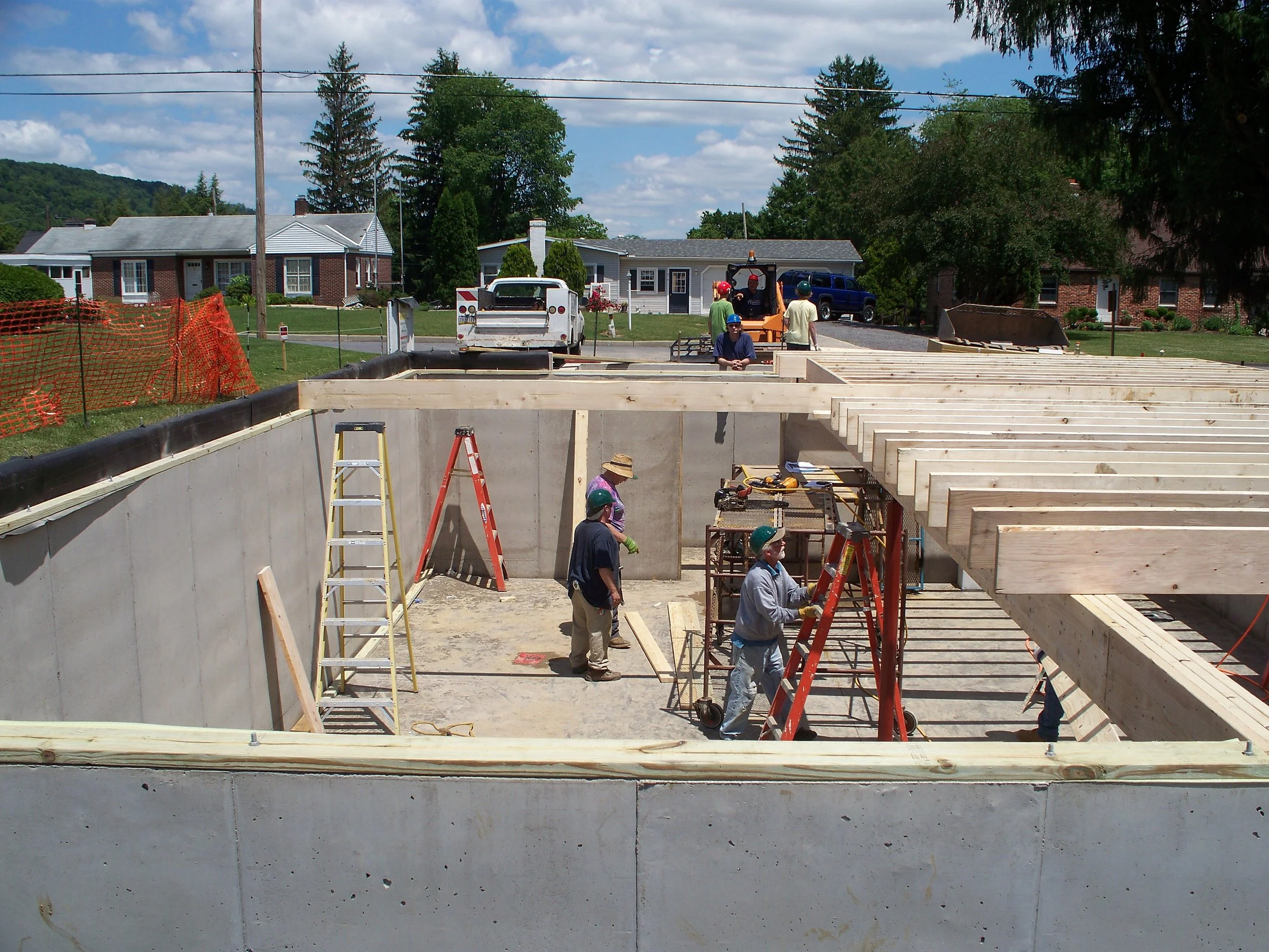 Construction workers building the foundation of a house, with wooden beams and ladders, in a residential neighborhood on a sunny day.