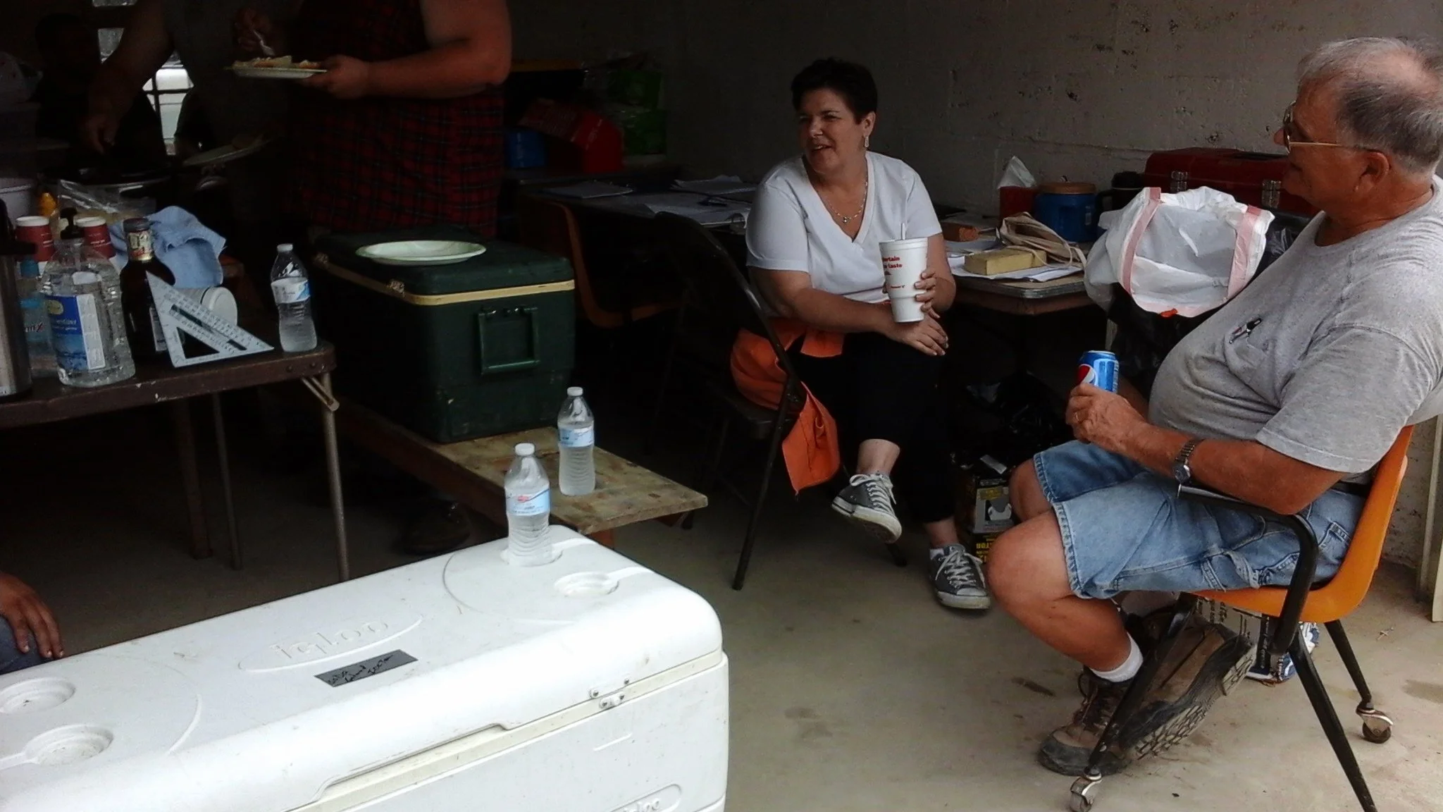Two people sitting in chairs, talking and holding drinks, surrounded by various bottles, water bottles, and cooler in a casual setting.