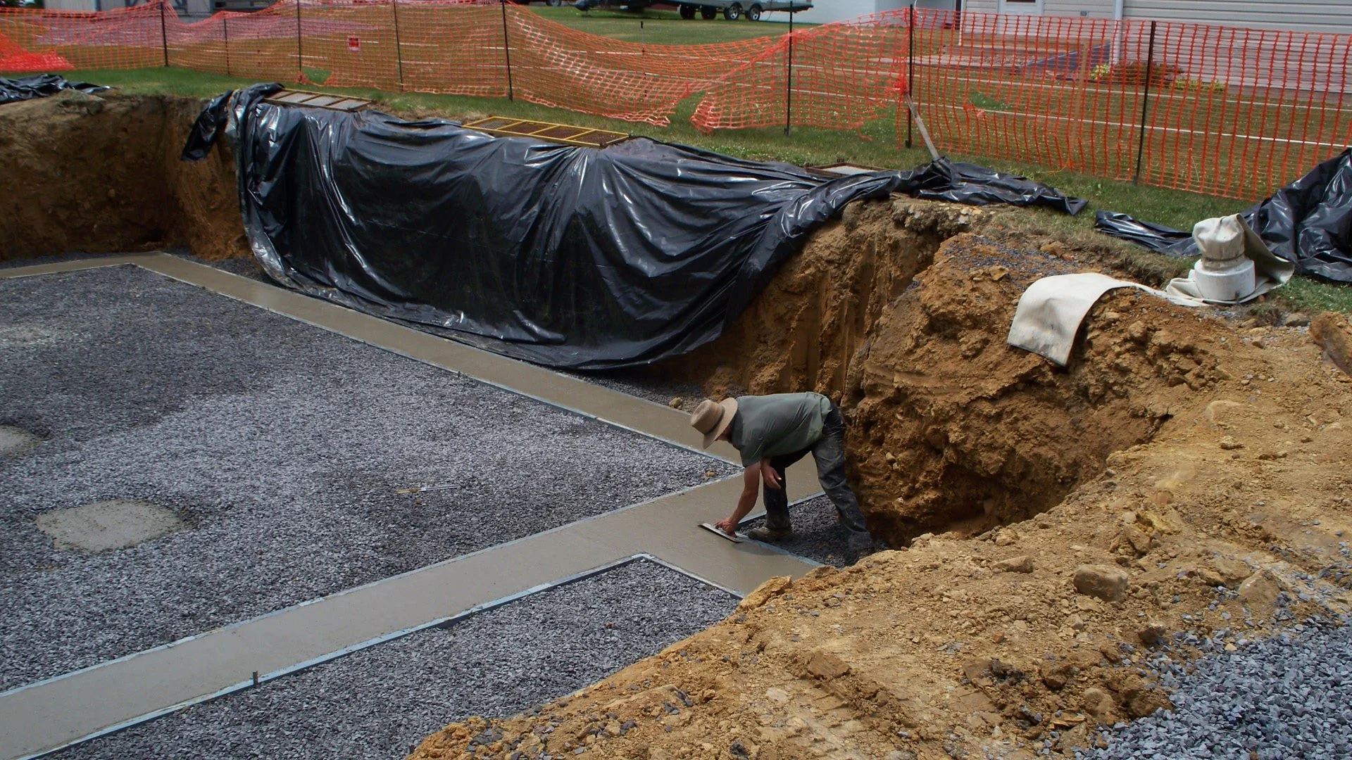 A construction worker is smoothing wet concrete in a foundation with a trowel at a construction site, surrounded by gravel, dirt, and orange safety fencing in the background.