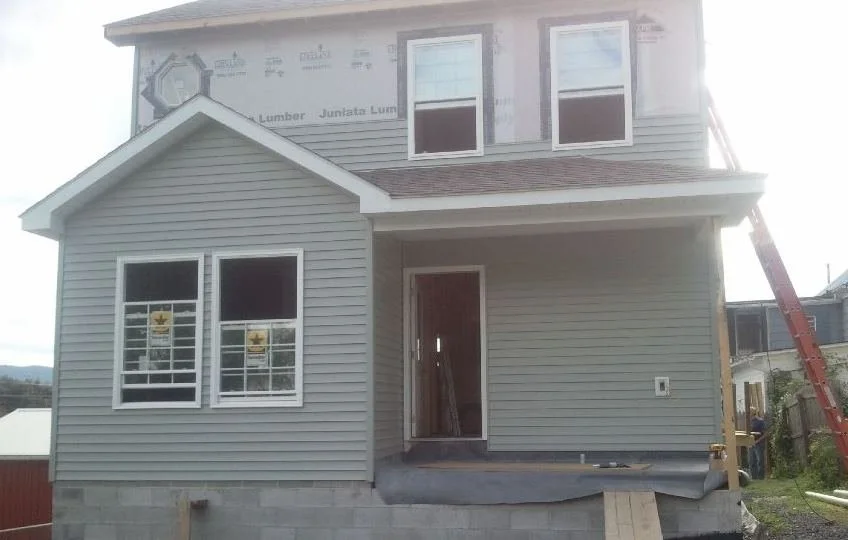 A house under construction with siding partially installed, a ladder leaning on the side, and construction materials visible.