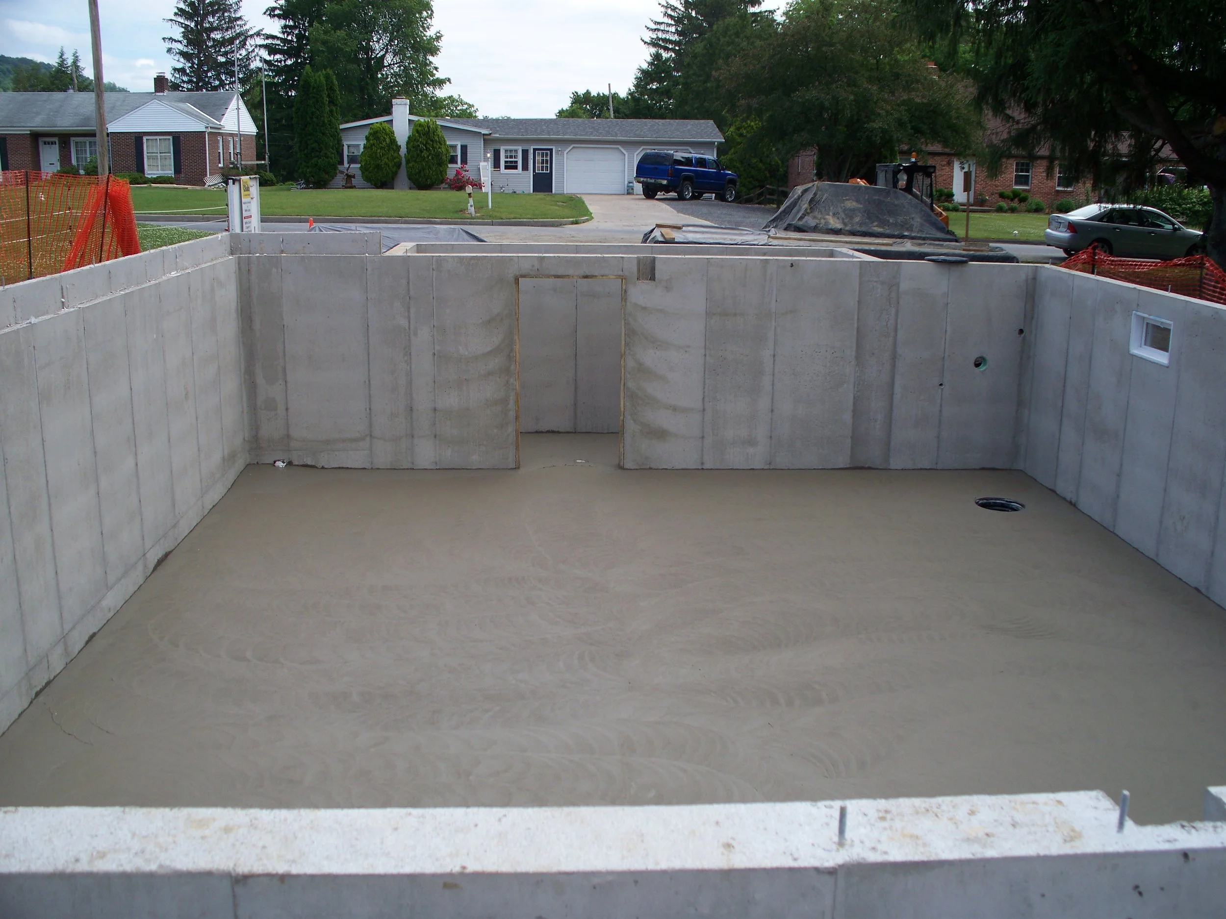 Construction site with a concrete basement foundation, light brown dirt floor, and concrete walls. Surrounding residential houses are visible in the background.