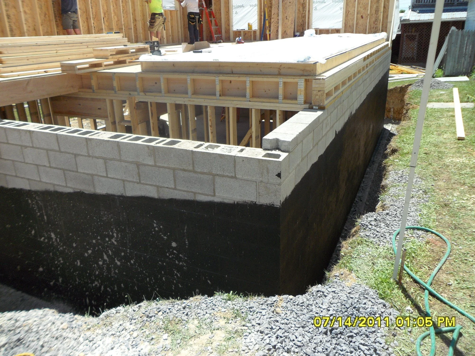 Construction site of a building with concrete blocks forming the foundation walls, wooden framing in progress, and construction workers working inside. The image shows a raised foundation with black waterproofing and gravel around the base.