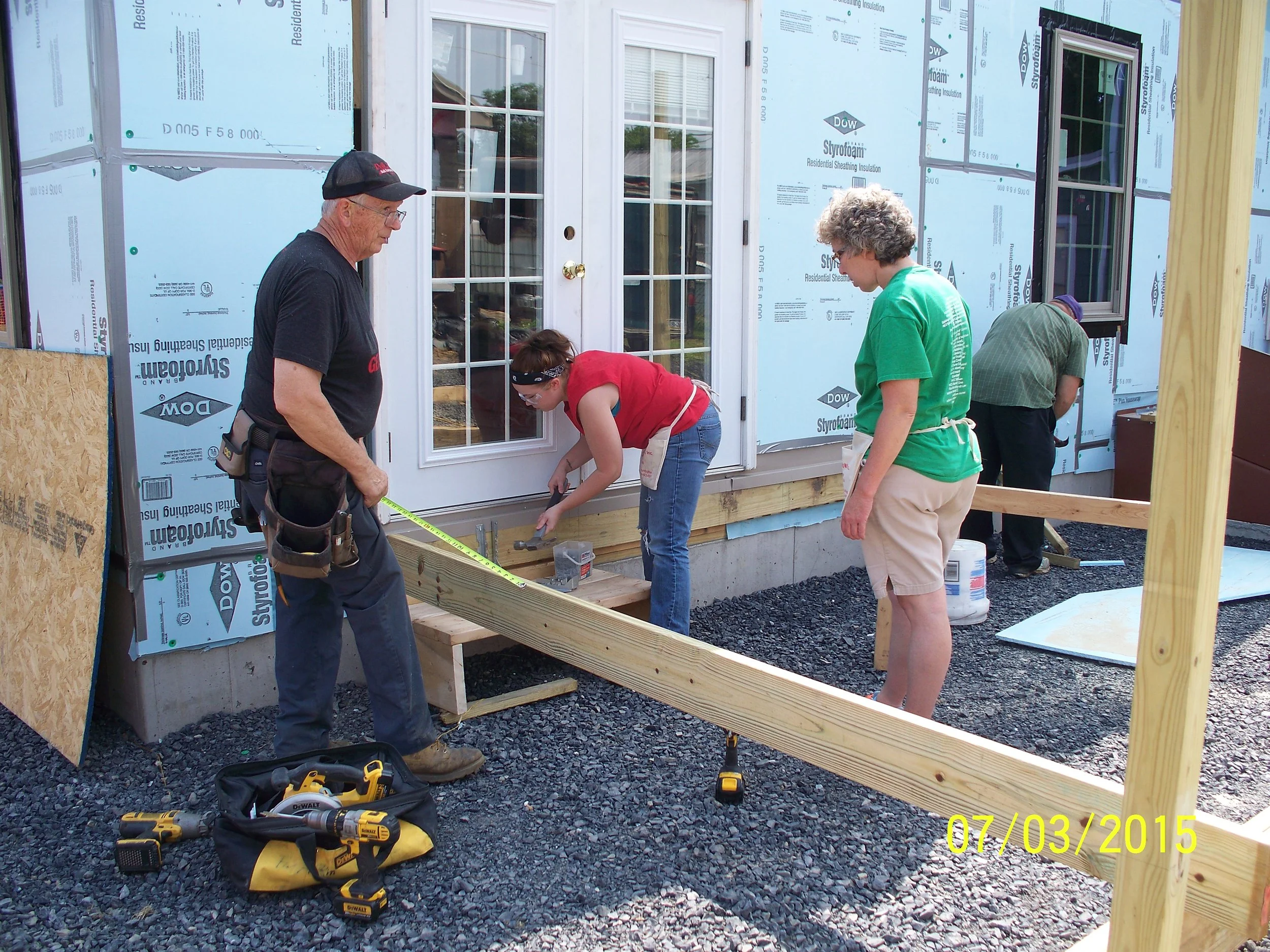 People working on a construction project outside a house, measuring and installing a wooden frame, with tools and equipment on the ground.