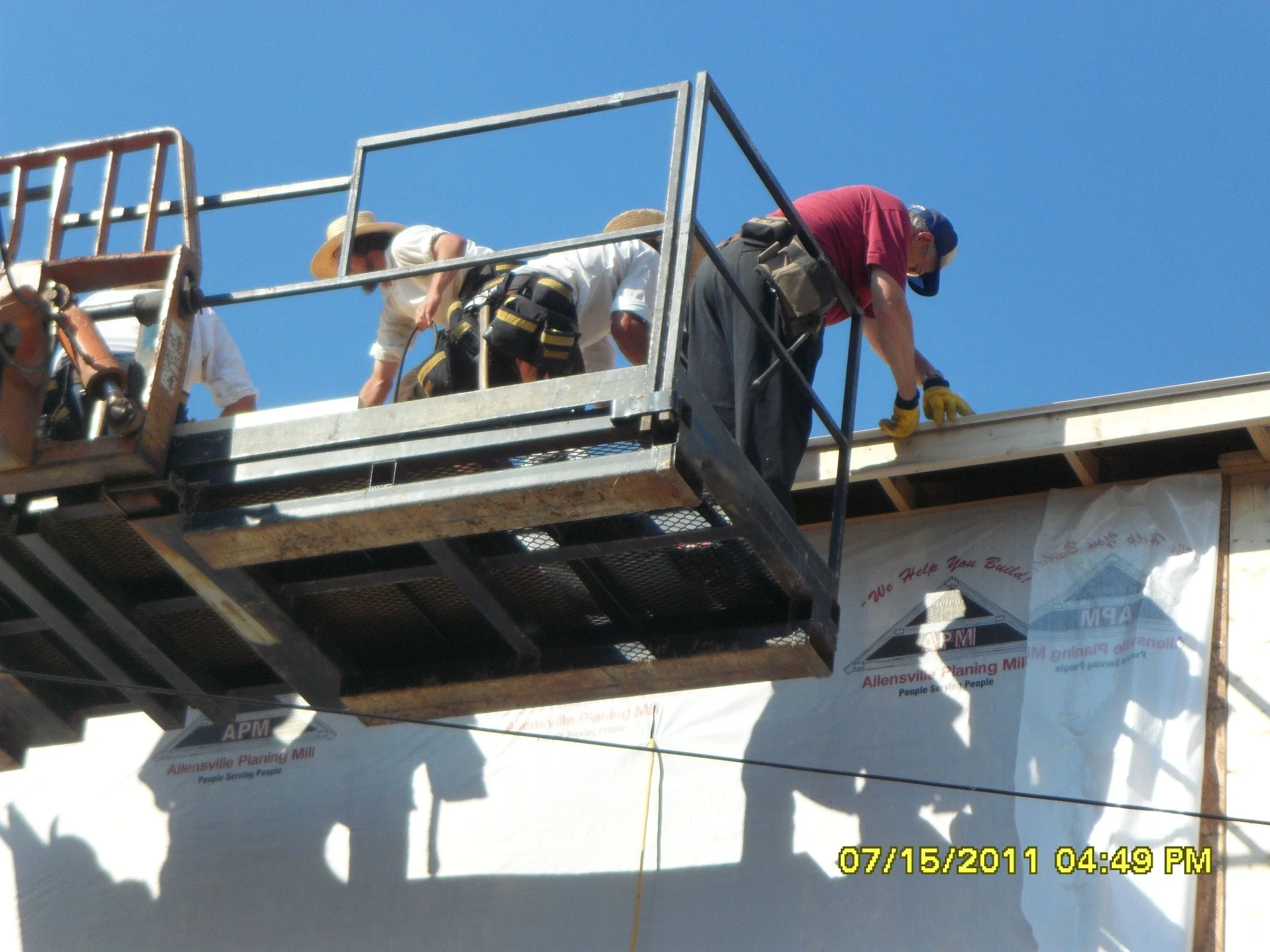 Construction workers installing a roof on a building, standing on an elevated platform with safety guardrails, under a blue sky.