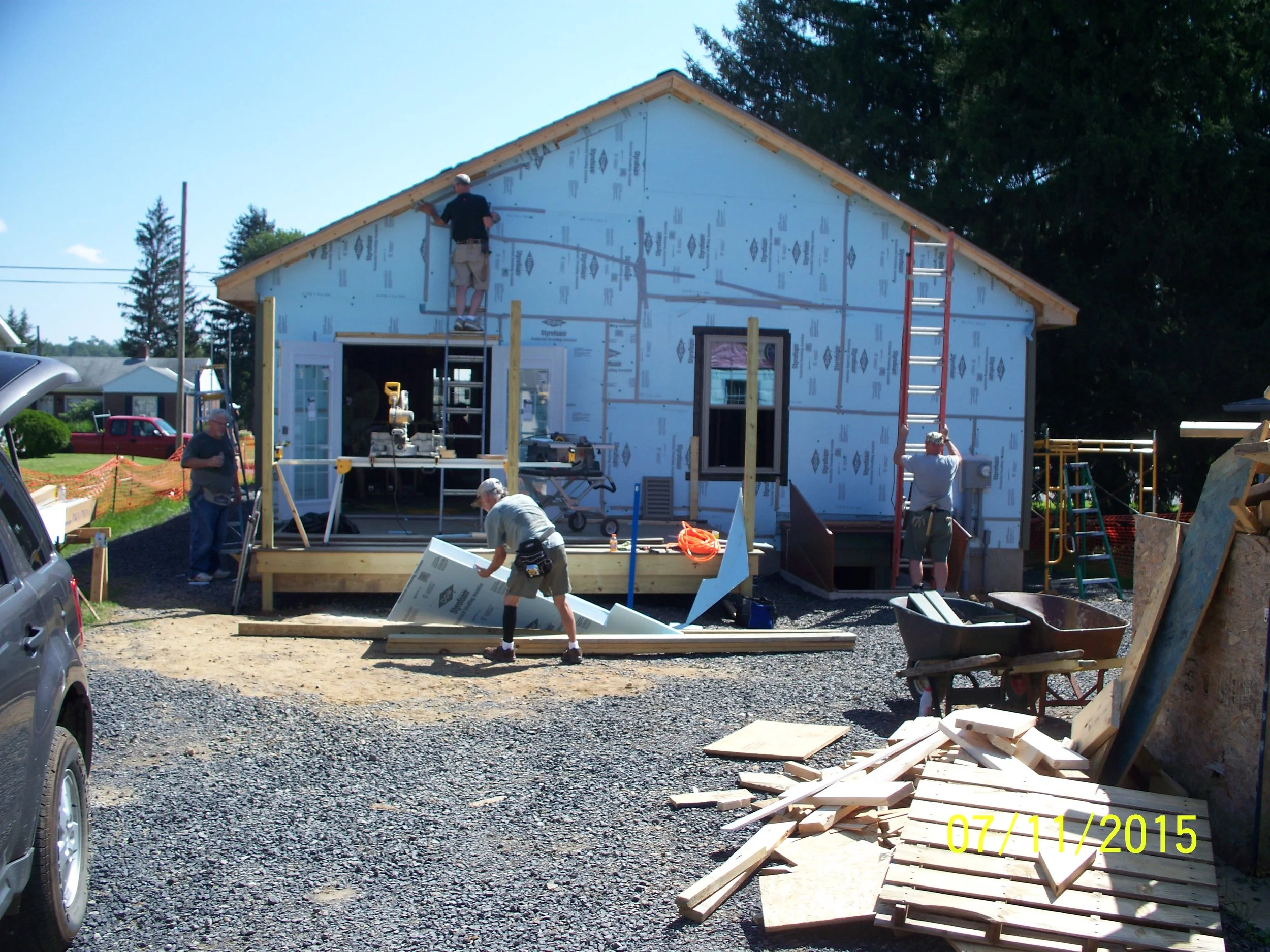 Construction workers building a house, with one on the roof installing siding, another on a ladder, and others working on the ground with tools and materials. The house is partially finished with blue sheathing and windows installed.