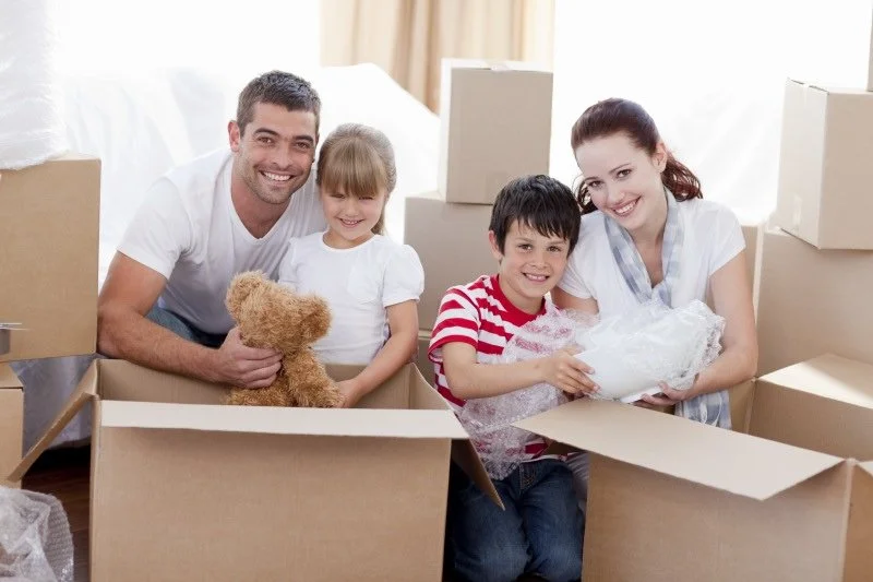 Happy family of four, including two children, preparing to move into a new home with packed boxes. The father is holding a teddy bear, and the mother and children are smiling with packed belongings.