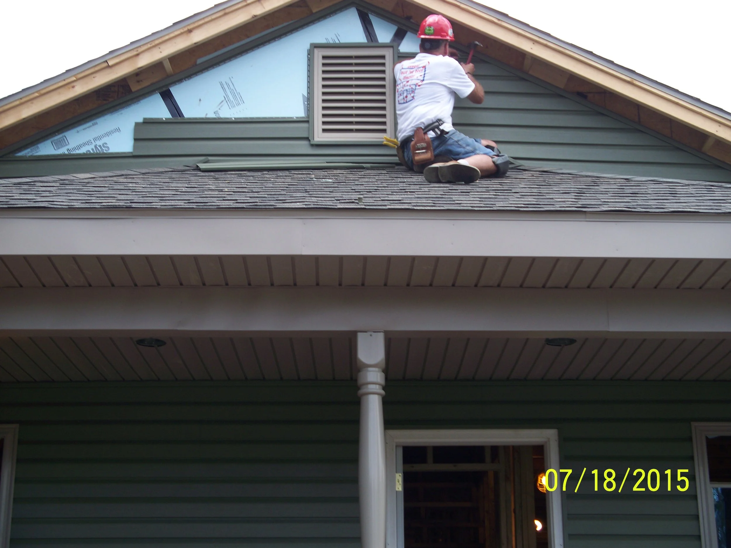 A construction worker on a roof installing or repairing a vent on a house. The worker is wearing a red hard hat, white shirt, and denim shorts, and is kneeling on the shingles.