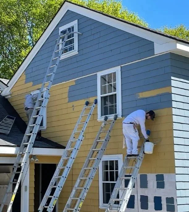 Two painters are working on painting the exterior of a house, using ladders to reach different sections of the upper walls. The house features blue and yellow sections with white window frames.