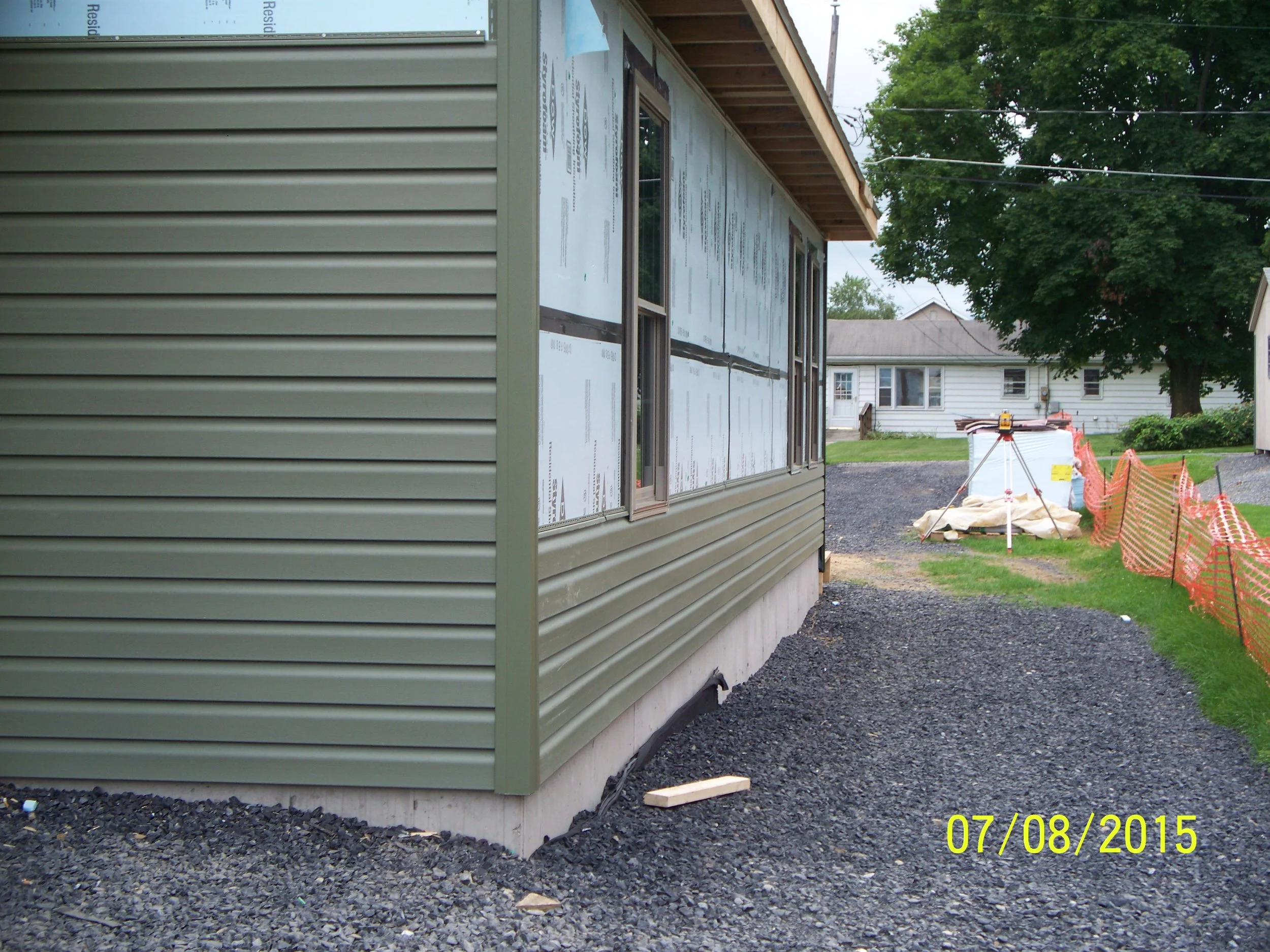 Side view of a house under construction with beige vinyl siding, gravel pathway, and orange safety fencing.