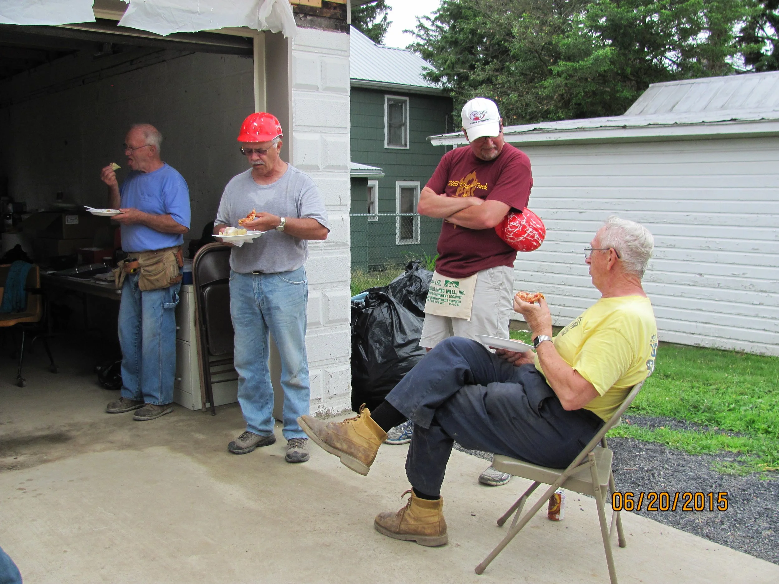 Four men are gathered outside near a garage and a white building. Three men are standing, eating pizza, while one man is sitting on a folding chair, holding pizza and a plate. The sitting man and the man standing to his right are older, with white ha