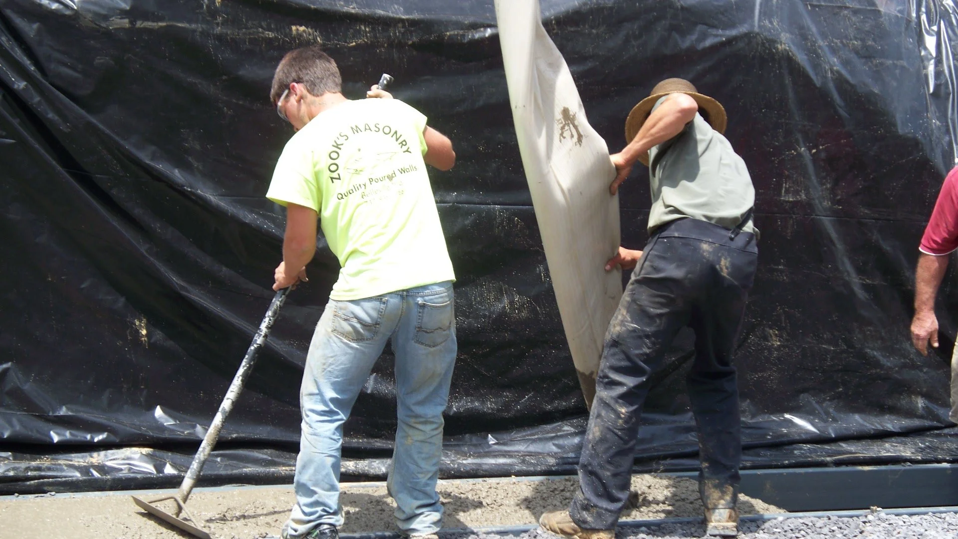 Two workers pouring concrete and leveling it with a tool while standing on a gravel surface next to a black tarp backdrop.