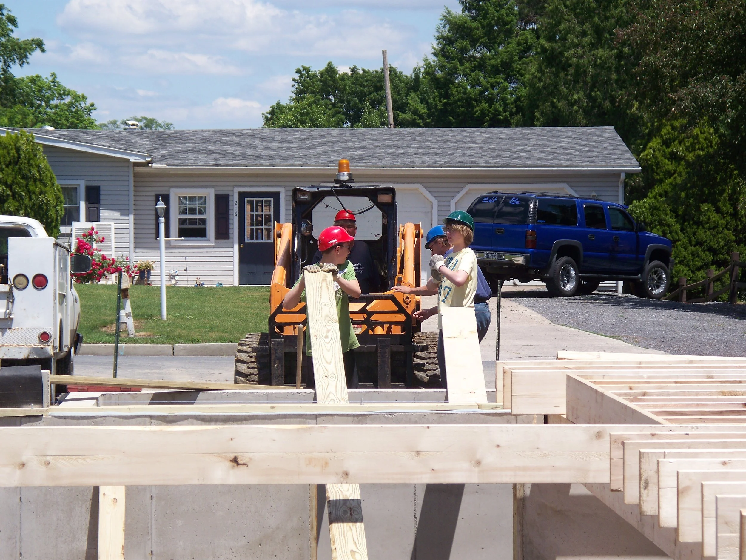 Children wearing helmets working on a construction site with a small backhoe, wooden beams, and a house in the background.