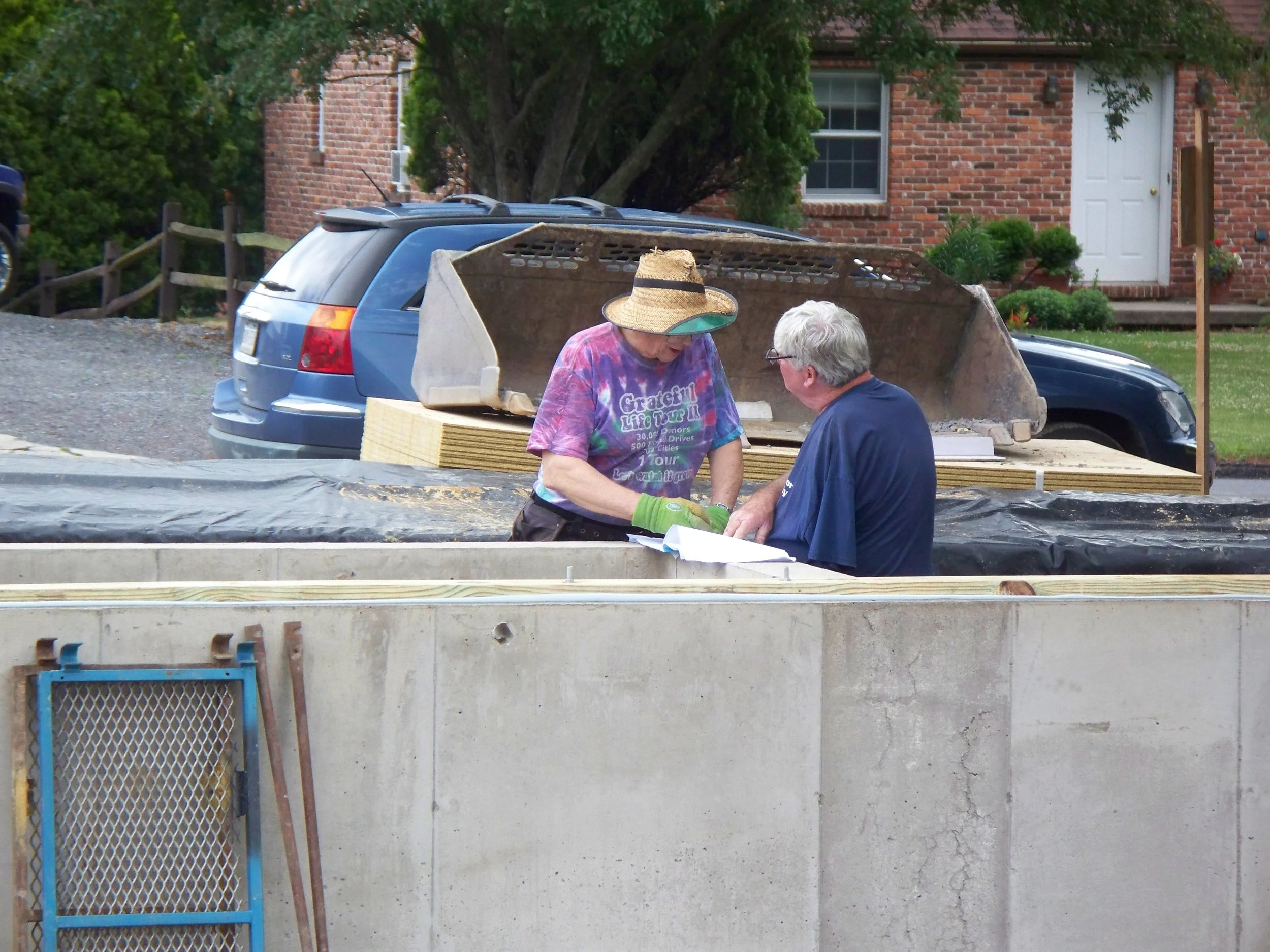 Two people on a construction site examining plans, with a concrete wall and construction materials in the background.