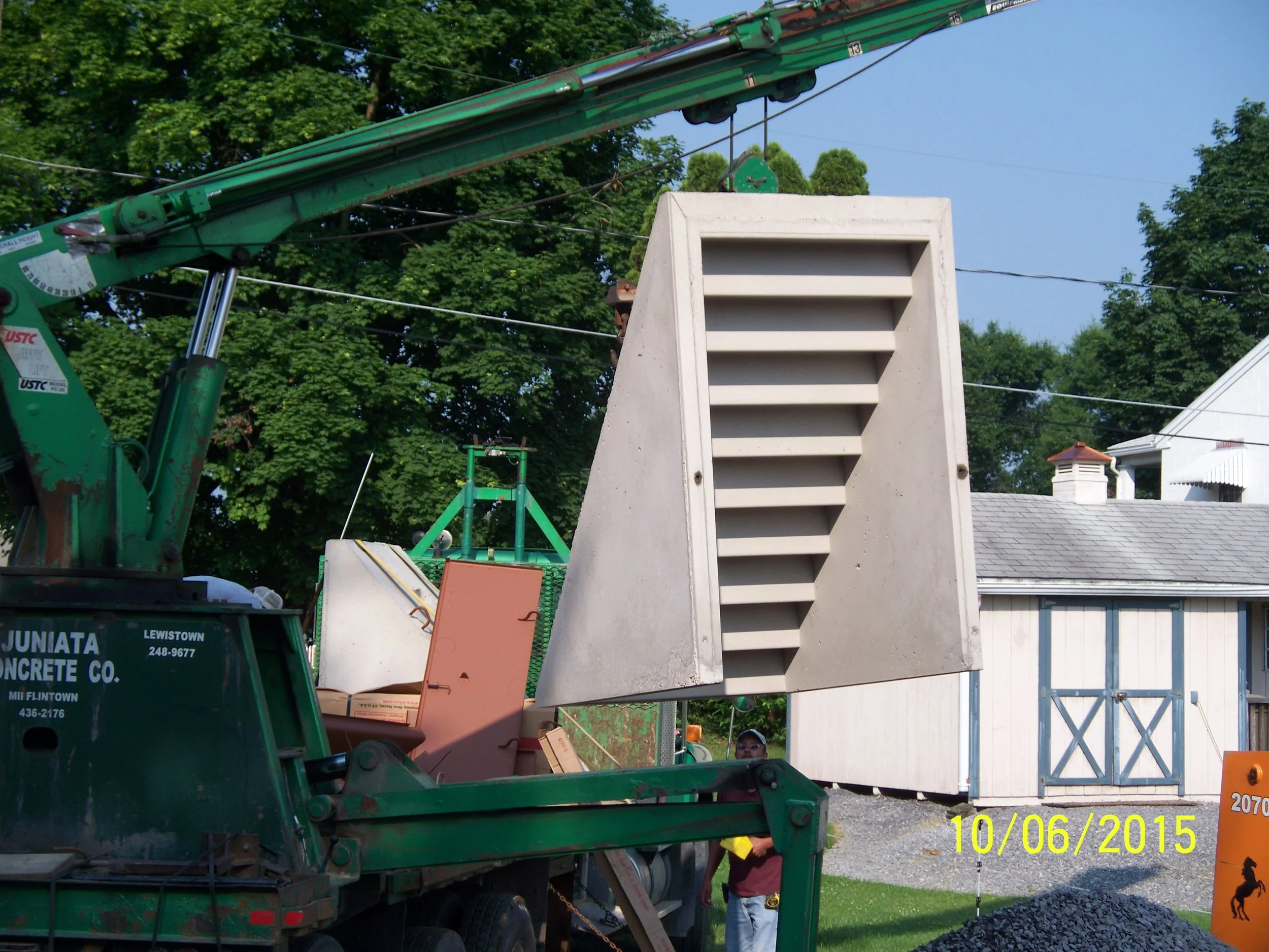 A green crane lifting a large concrete air vent with horizontal slats into the air at a construction site. There are trees, a white building with blue doors, and construction equipment in the background. The photo is dated October 6, 2015.