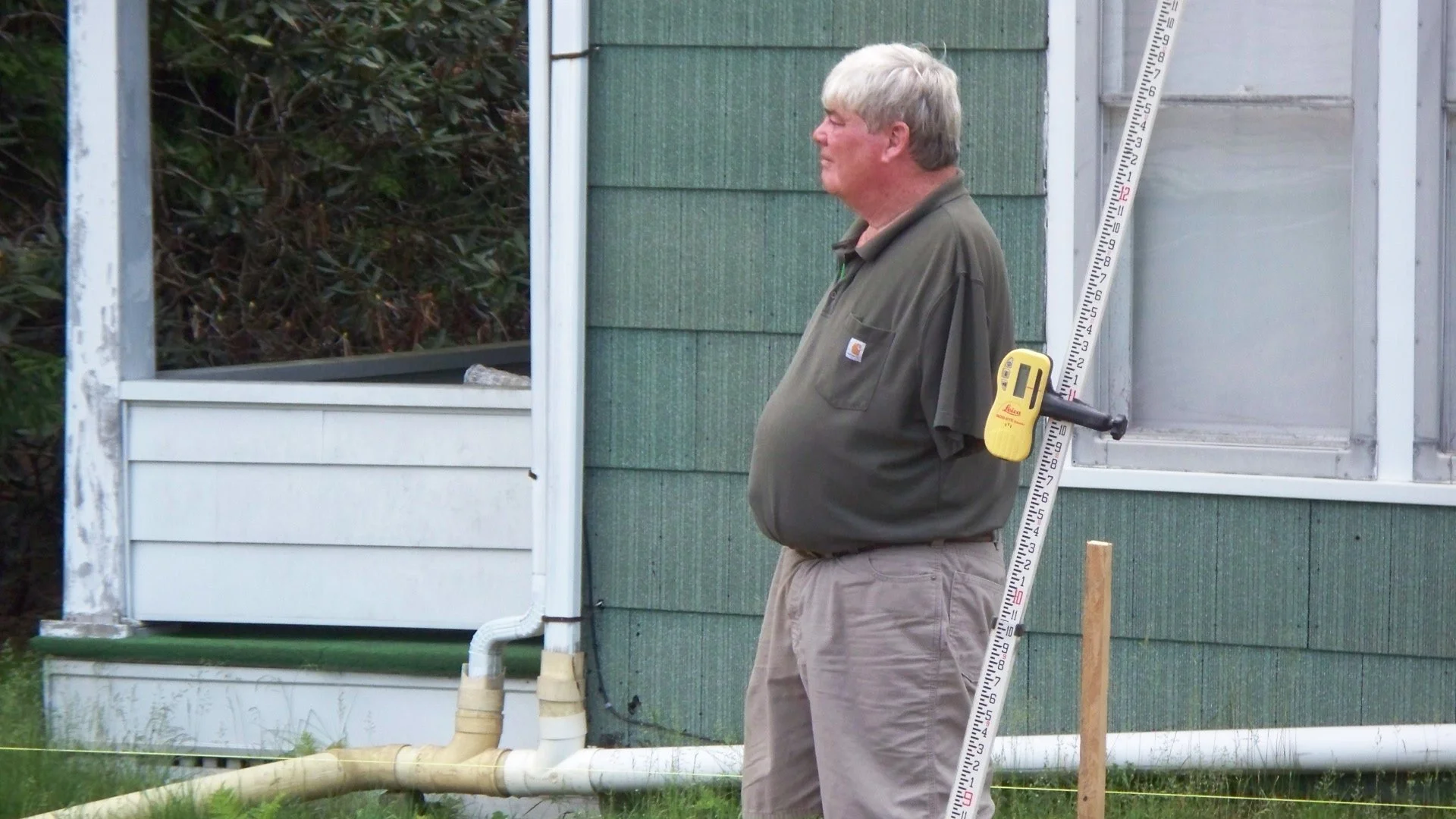 A man with gray hair wearing a brown Carhartt shirt standing outdoors beside a green wooden house with pipes and measuring equipment attached to the house.