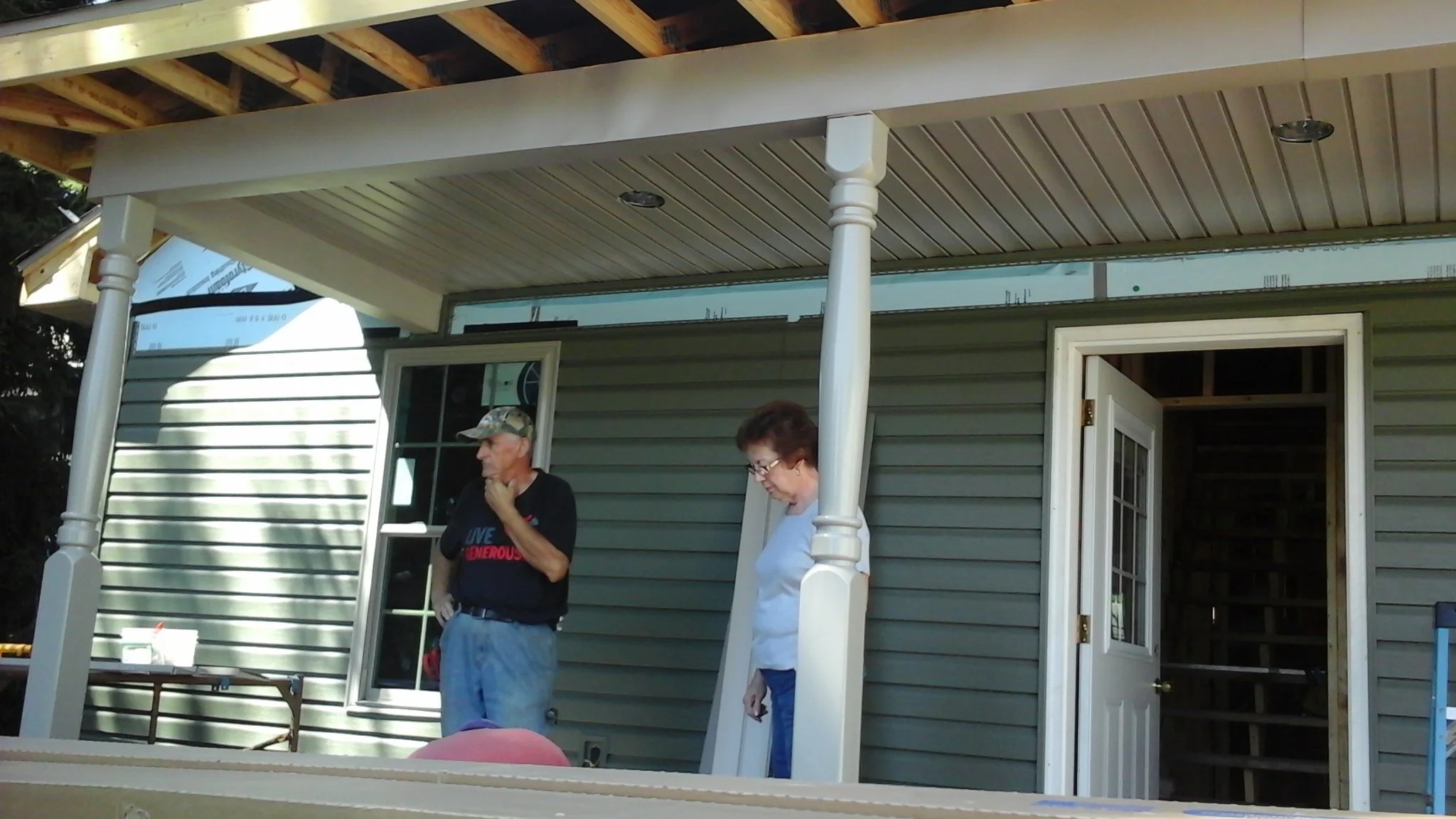 Two people standing on a porch of a house under construction, with unfinished exterior walls and an open door leading inside. One man wears a black T-shirt and camouflage cap, and a woman wears glasses and a white top.