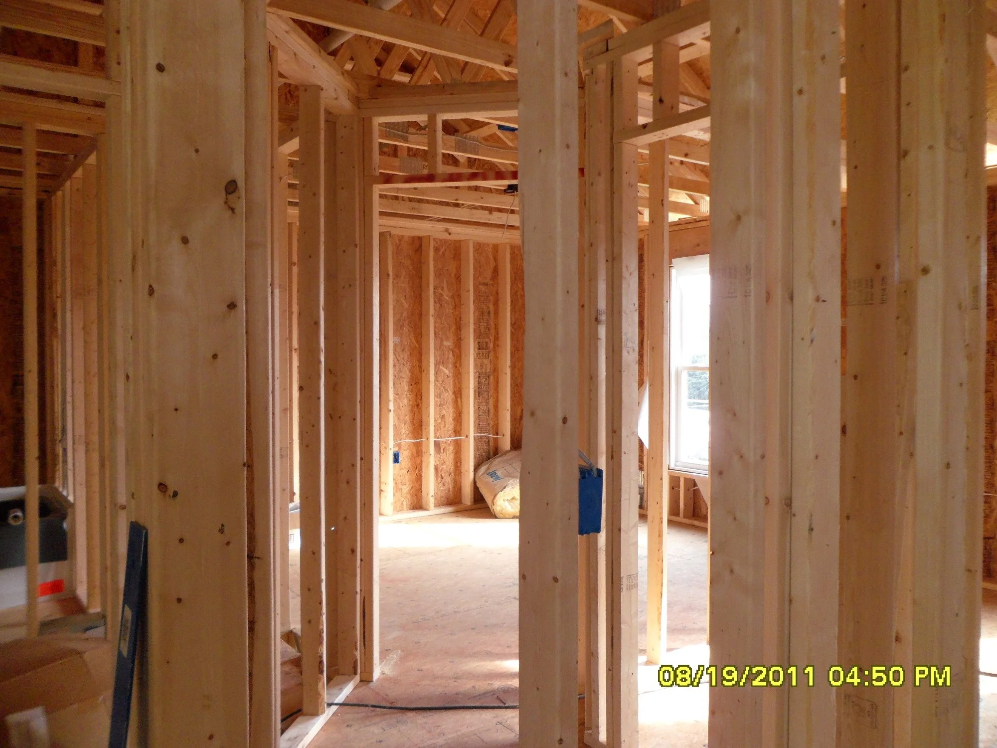 Interior of a house under construction with exposed wooden studs and framing, with a window on the right allowing sunlight inside.