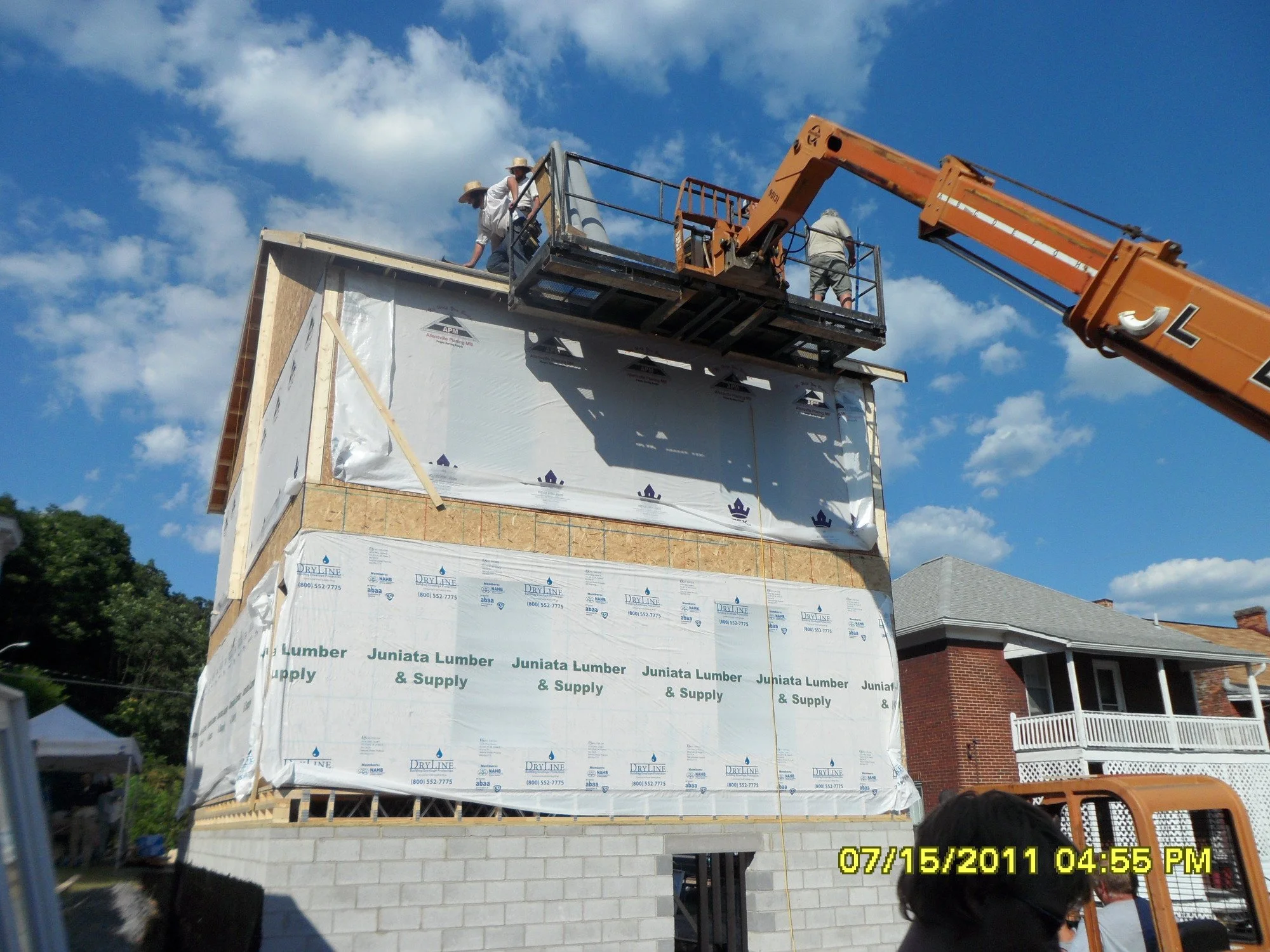 Construction workers on a lift working on the upper part of a house under construction, with a clear blue sky and some clouds in the background.