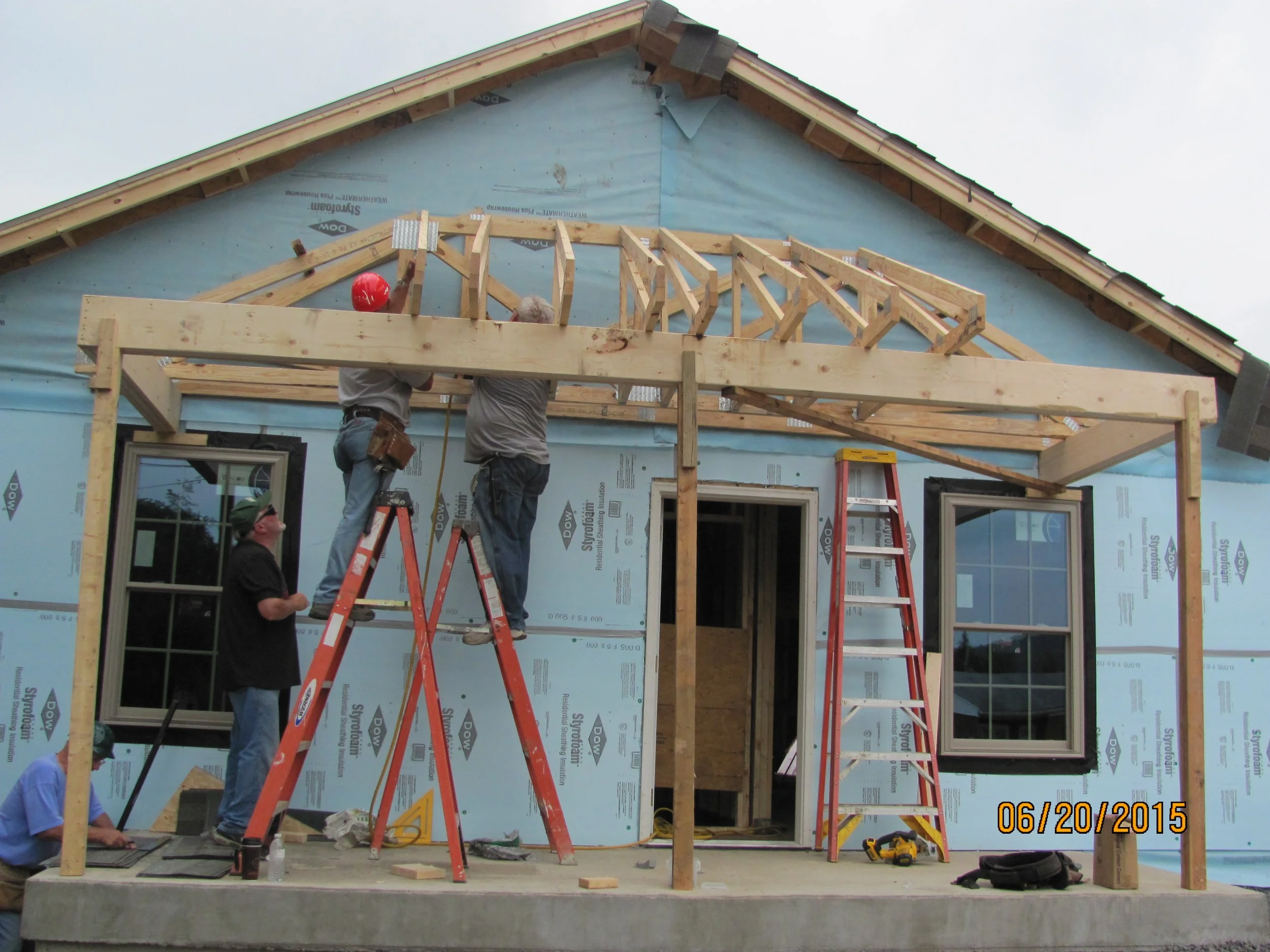 Construction workers building the wooden frame/orientation of a house's porch or roof extension, with ladders and construction tools visible.