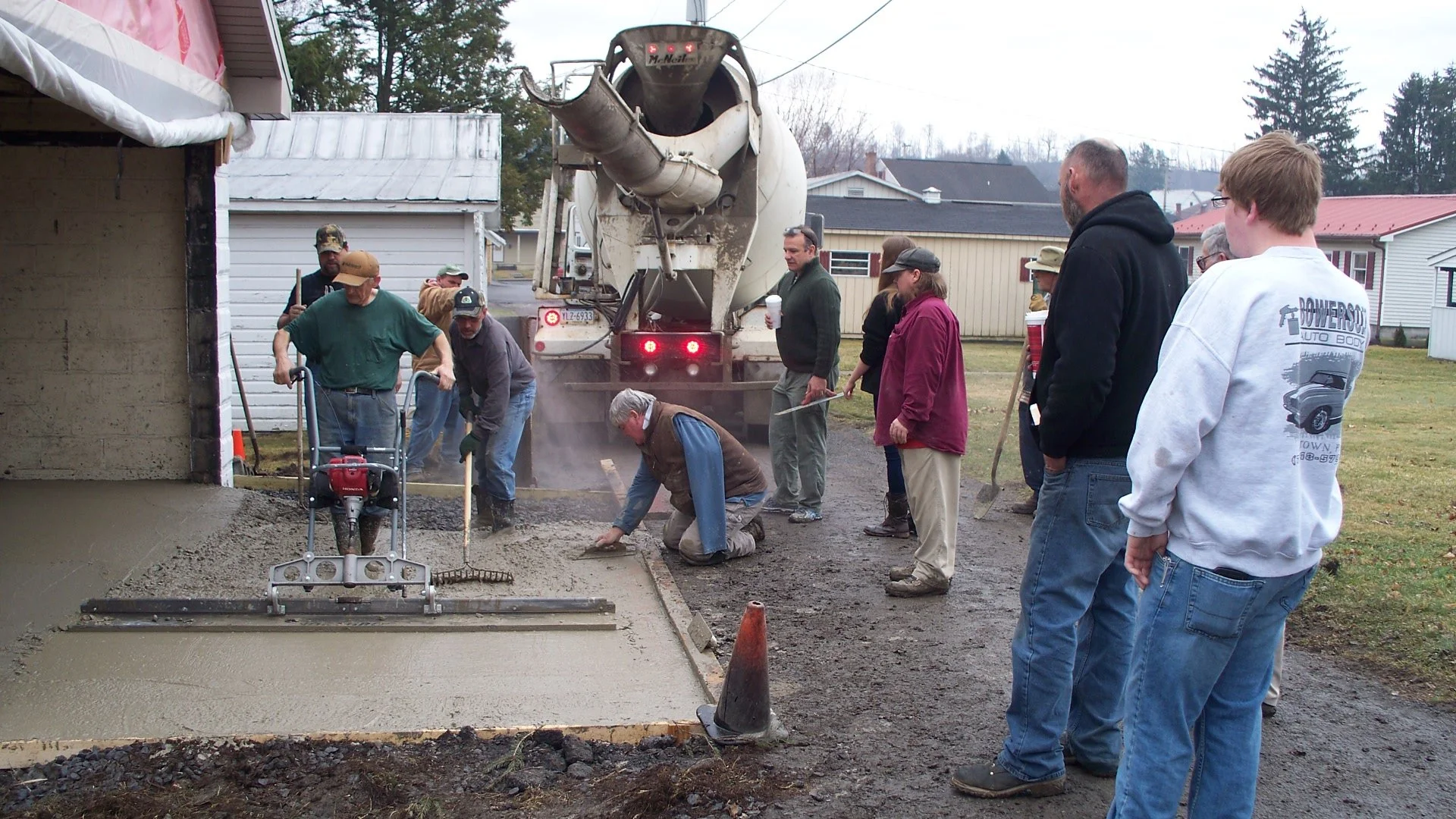 Group of workers and onlookers pouring and smoothing concrete for a sidewalk or driveway outside a house.