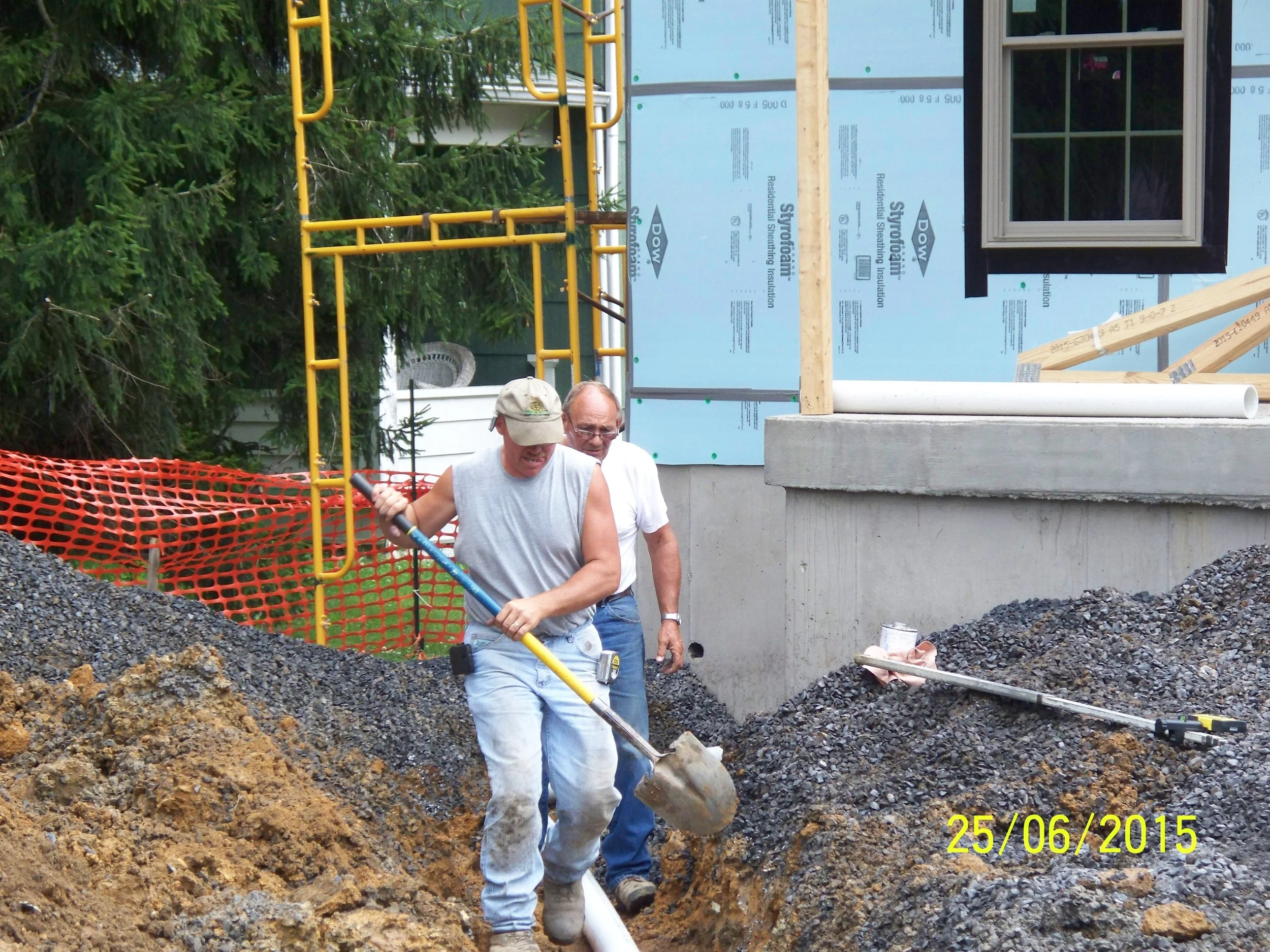 Two men working on a construction site, one shoveling gravel and the other standing behind him. There are building materials and a window frame visible, with a safety fence in the background, and trees nearby.