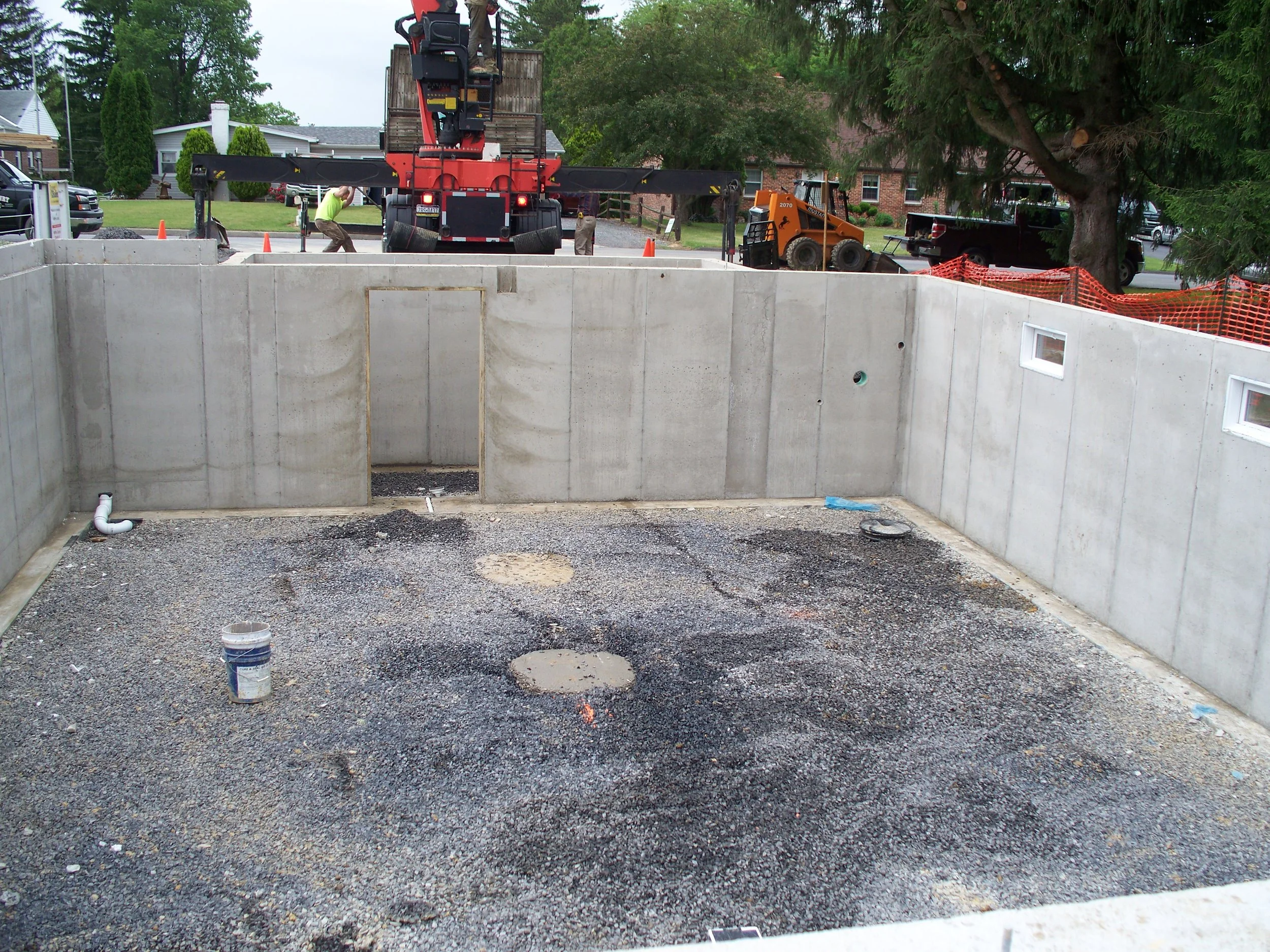 Construction site of a building foundation with concrete walls and gravel ground, with construction equipment and workers visible in the background.
