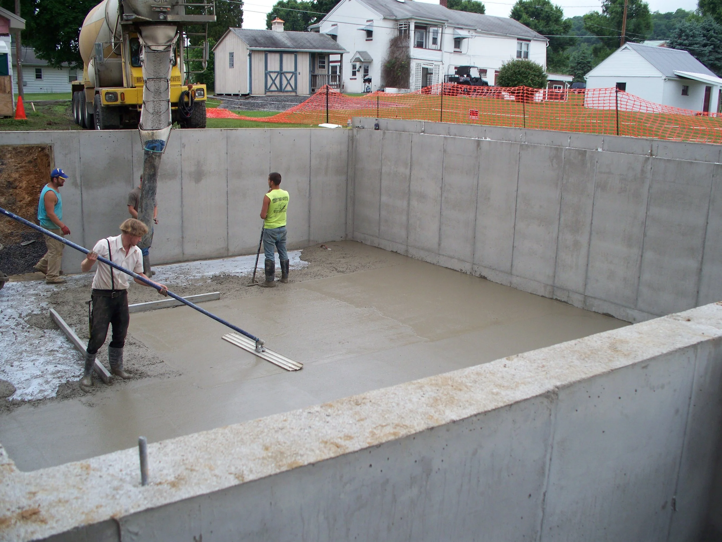 Construction site with workers pouring and leveling concrete in a rectangular foundation, surrounded by a concrete wall, with houses and trees in the background.
