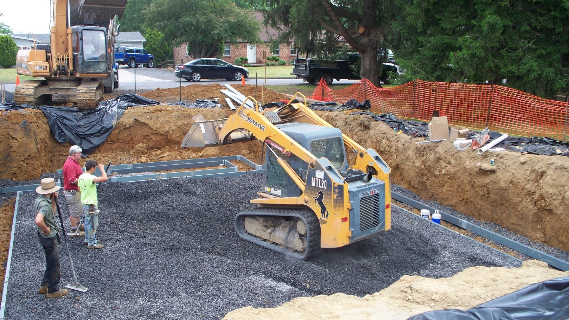Construction site with a small bulldozer leveling gravel, three workers inspecting, large excavator in the background, orange safety fencing, residential houses and trees nearby.