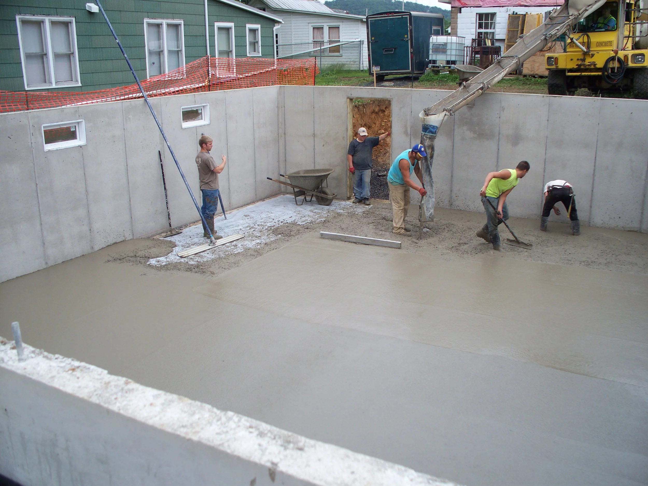 Construction workers are pouring and leveling concrete in a basement or foundation of a building, with construction equipment and tools visible.