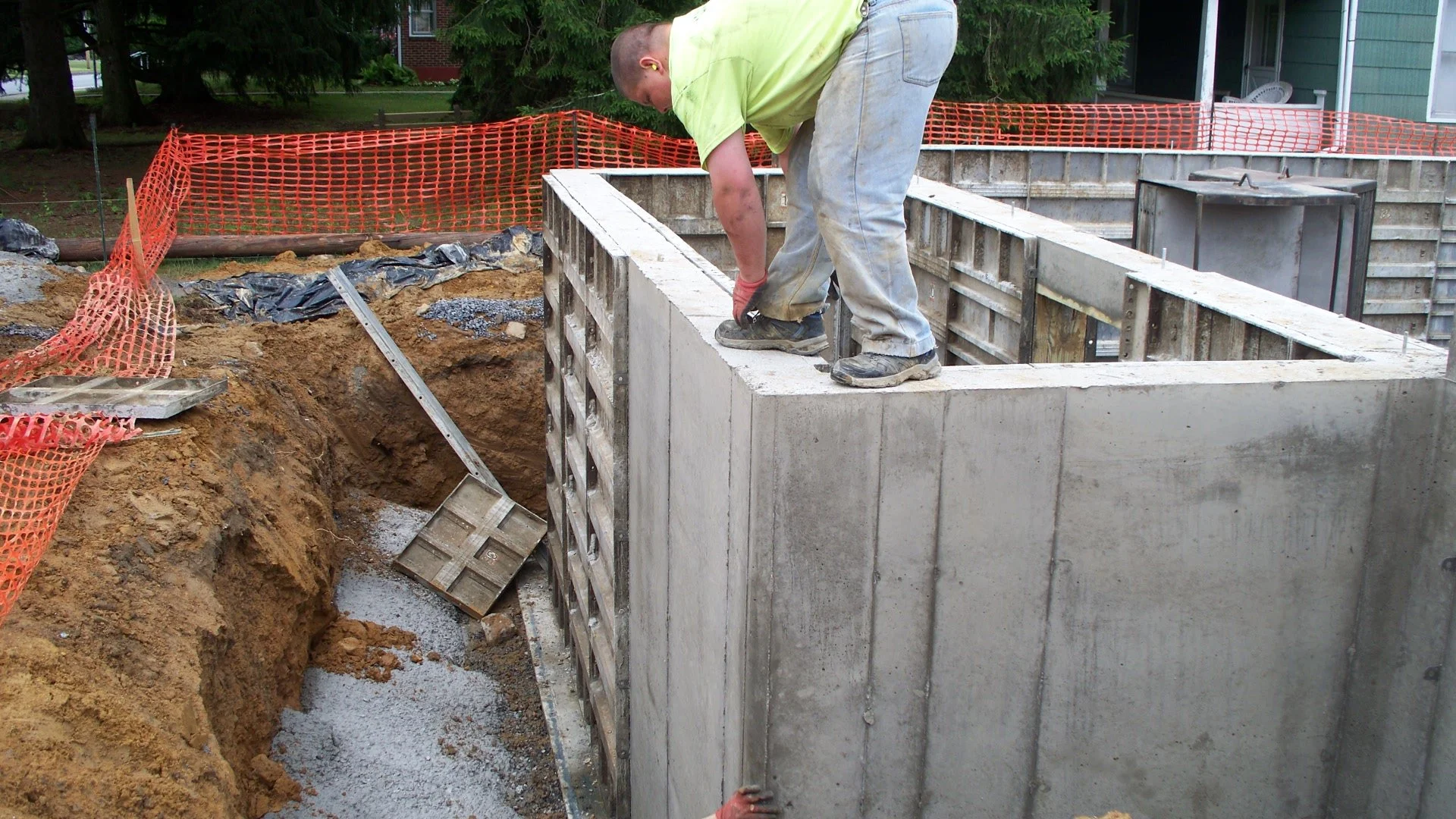 A construction worker installing a concrete wall at a construction site, surrounded by dirt and orange safety fencing.