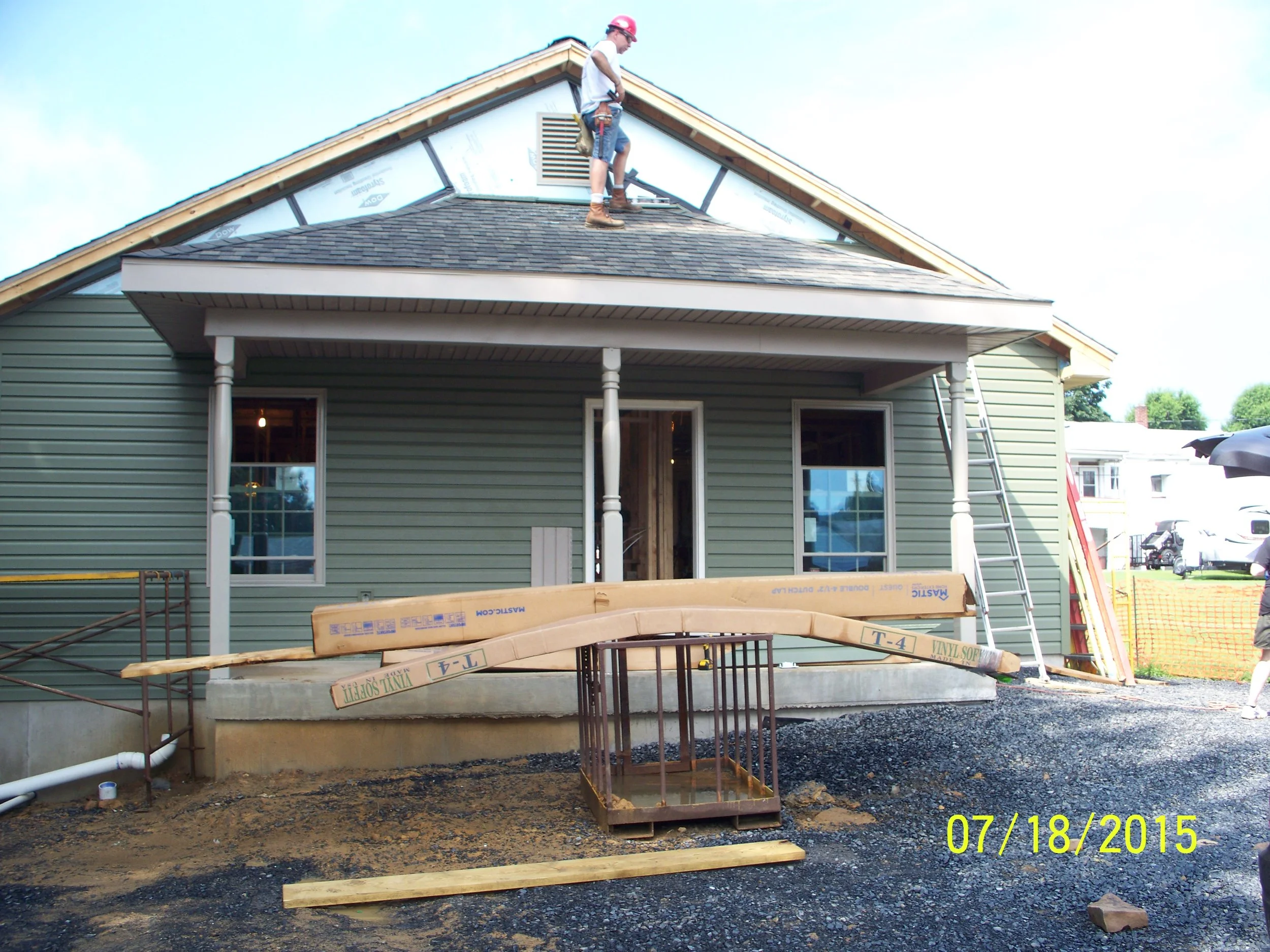Construction workers working on the roof of a house, with tools and building materials around. The house has green siding and white trim, with a porch supported by columns.