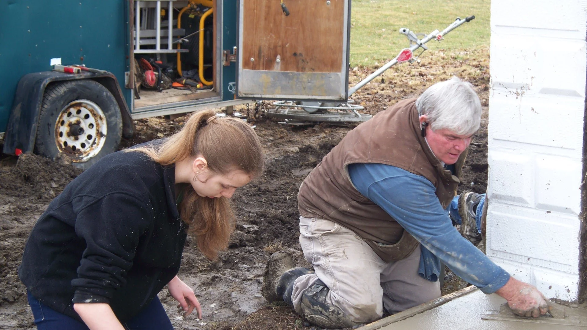 Two people are working on construction, laying a concrete foundation or wall next to a building. The older man is smoothing the concrete with a trowel, while the young woman is observing or assisting. They are outdoors on a muddy ground with a traile