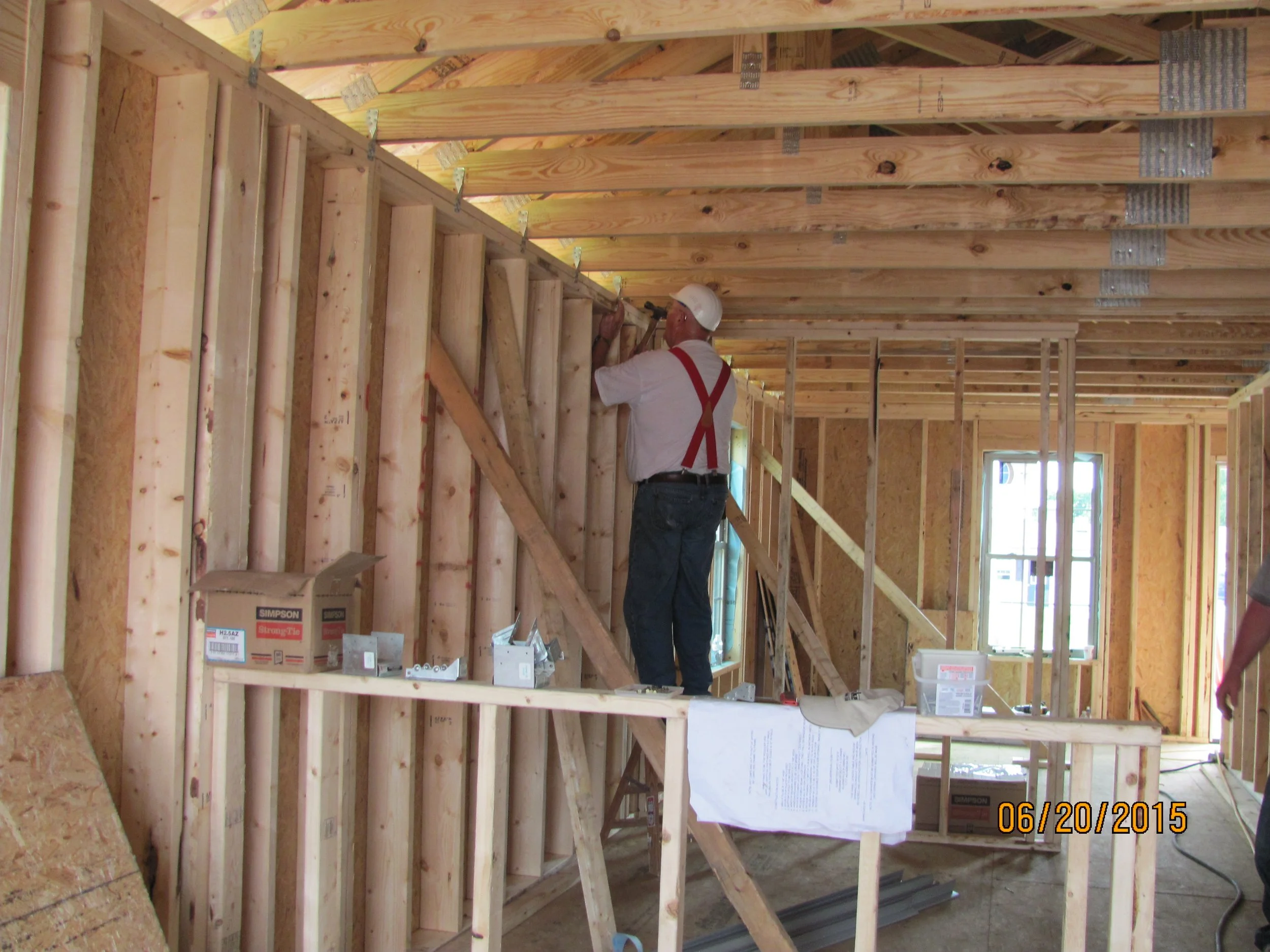 A construction worker wearing a white helmet and red suspenders is installing or inspecting wooden wall framing inside a house under construction, with windows and unfinished walls visible.