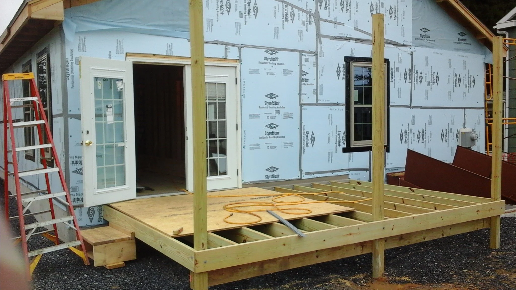 Front view of a house under construction with an attached deck framework. The house has a door and a window, with exterior insulation visible. A red ladder is on the left side, and construction materials and tools are present around.