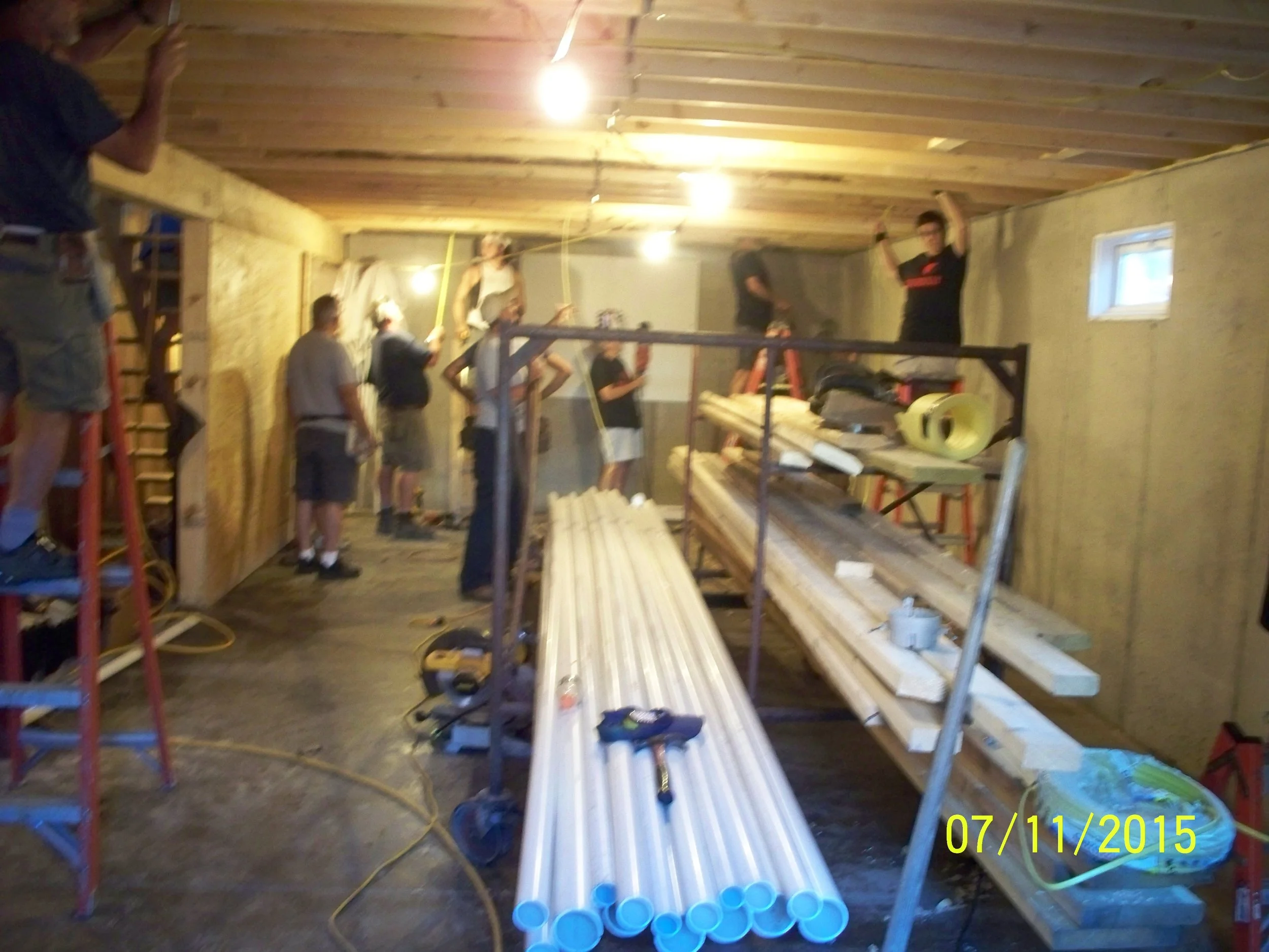 People working on constructing or renovating an interior space, with construction materials, tools, ladders, and pipes visible, inside a room with unfinished walls and ceiling.