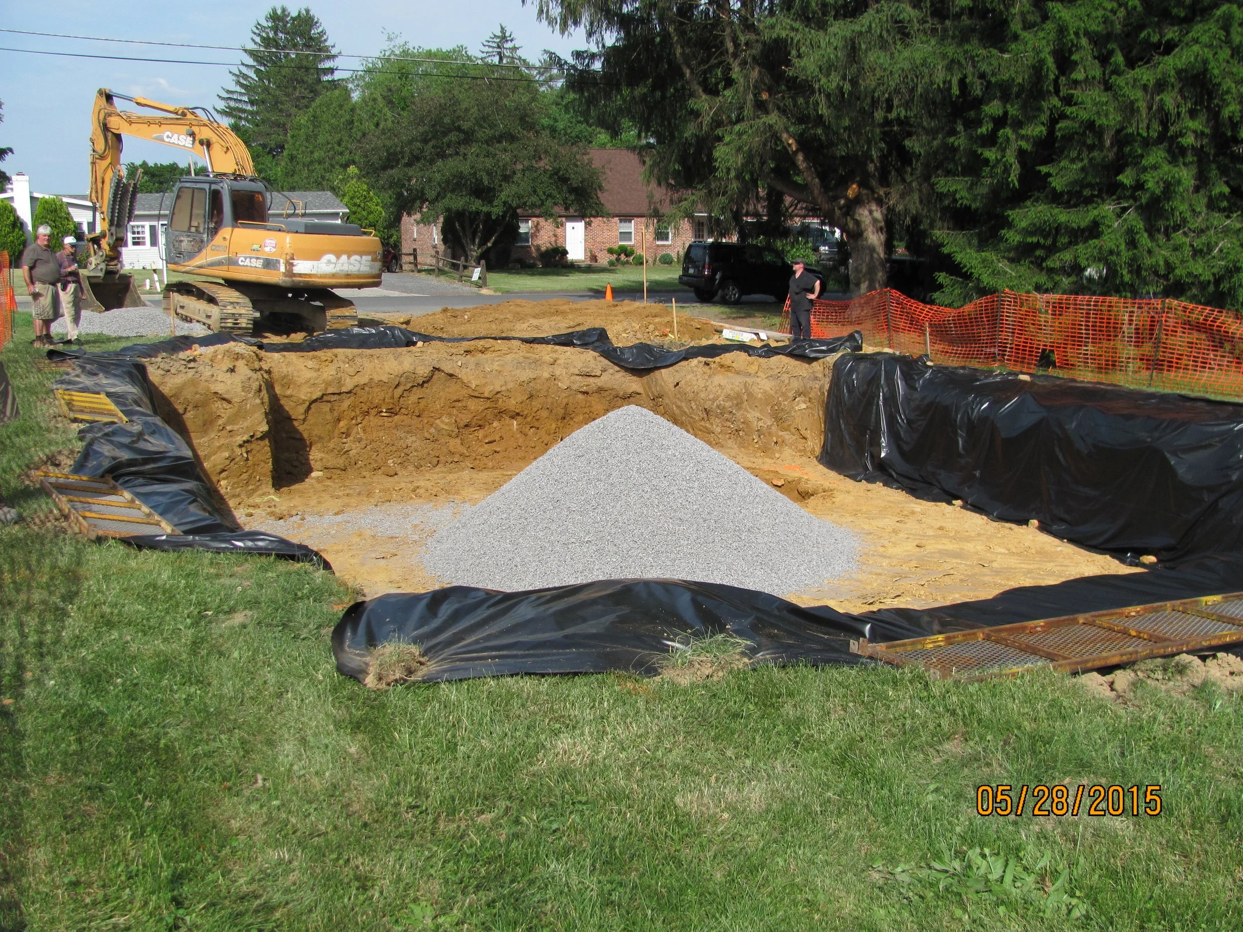 construction site with an excavator digging a hole, black plastic sheeting lining the edges, and a pile of gravel in the center, with people and trees in the background.