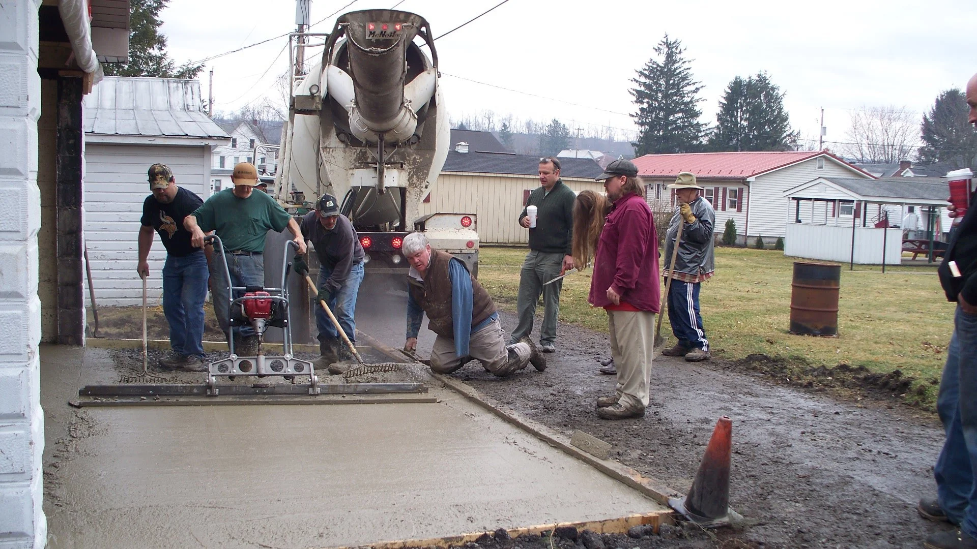 People working on pouring and leveling concrete for a sidewalk outside a building on a cloudy day; a concrete mixer truck is in the background.