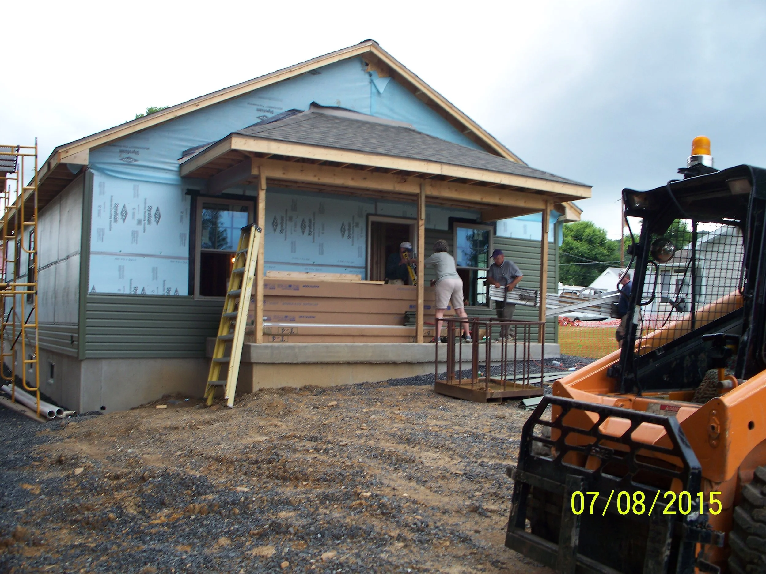 Construction site of a house with workers installing the front porch, surrounded by equipment and materials, with a partially finished exterior wall.