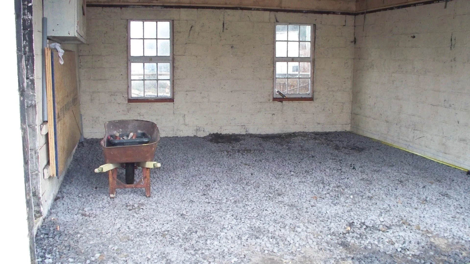 Interior of a room under construction with gravel floor, one wheelbarrow, and unfinished walls with two windows.