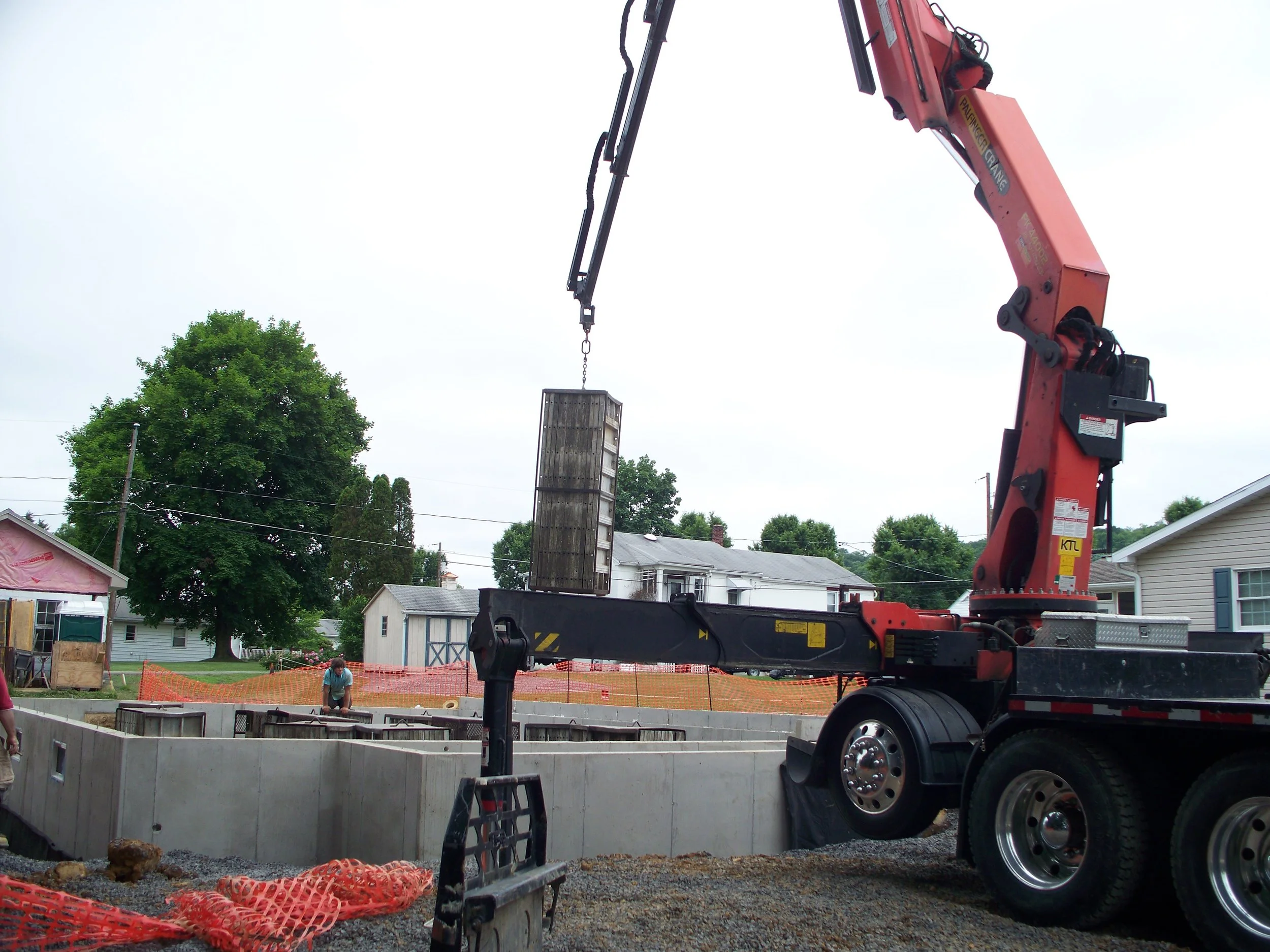 A construction site with a large red crane lifting a concrete form into place, surrounded by unpaved ground and residential houses in the background.