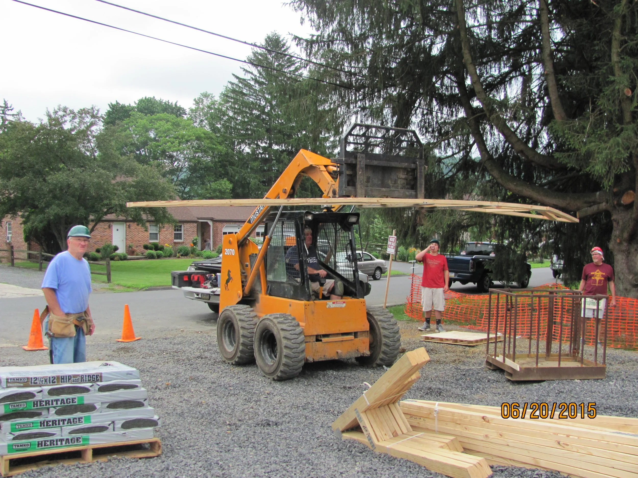 Construction site with a small forklift lifting a wooden platform, four men, some wearing safety hats, standing around, safety cones, stacked bags, and wooden materials on gravel ground surrounded by trees and houses.