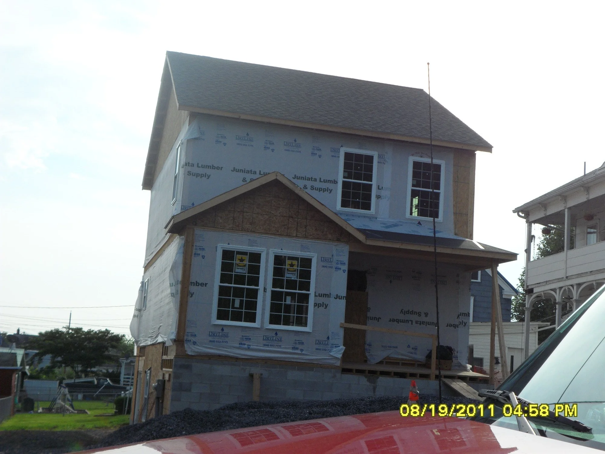 A two-story house under construction with exterior sheathing and windows installed, surrounded by construction materials and equipment, with neighboring homes visible in the background.