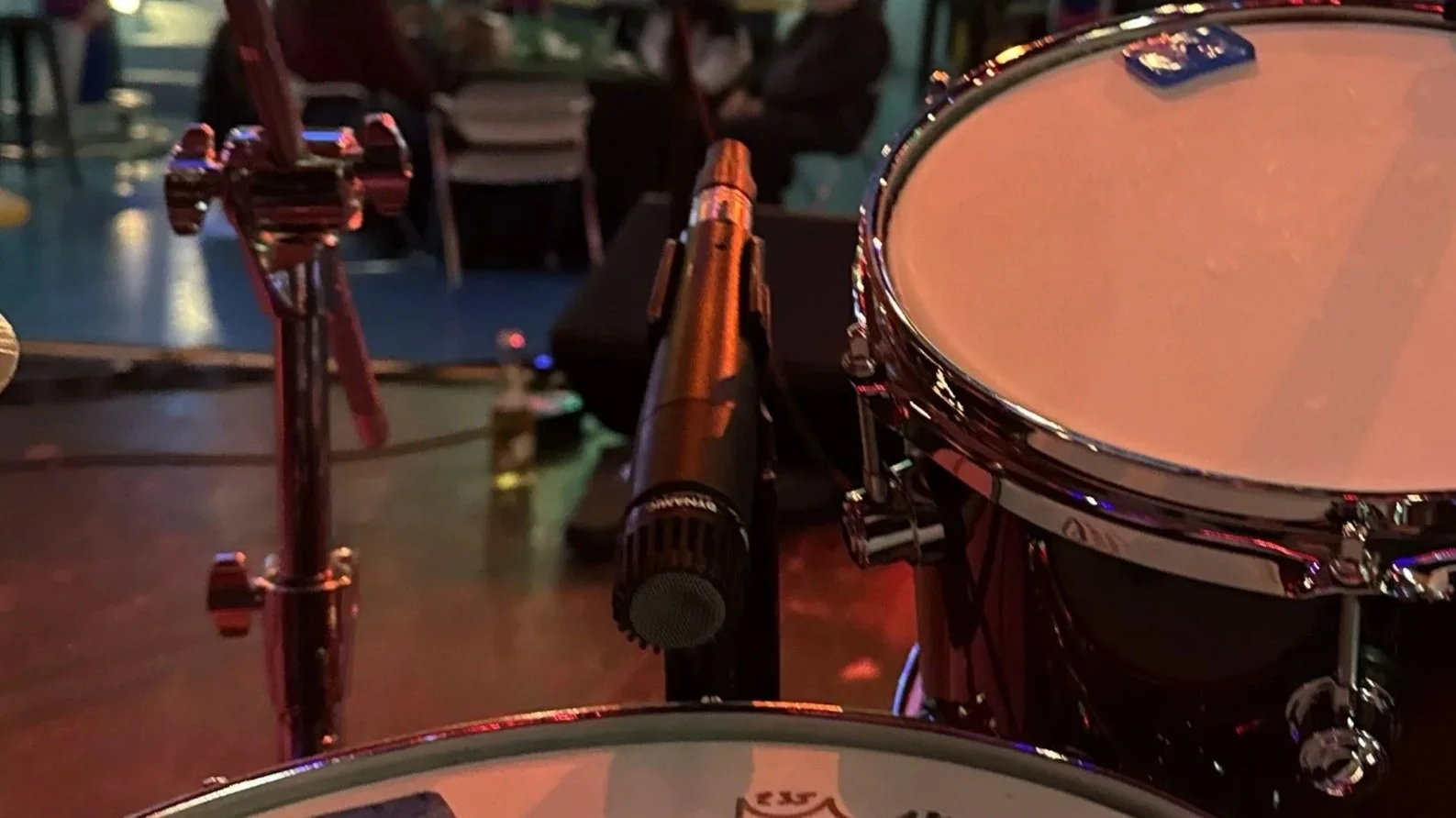Close-up view of a drum set with a microphone positioned in front of a snare drum, and a music stand in the background, in a dimly lit setting.