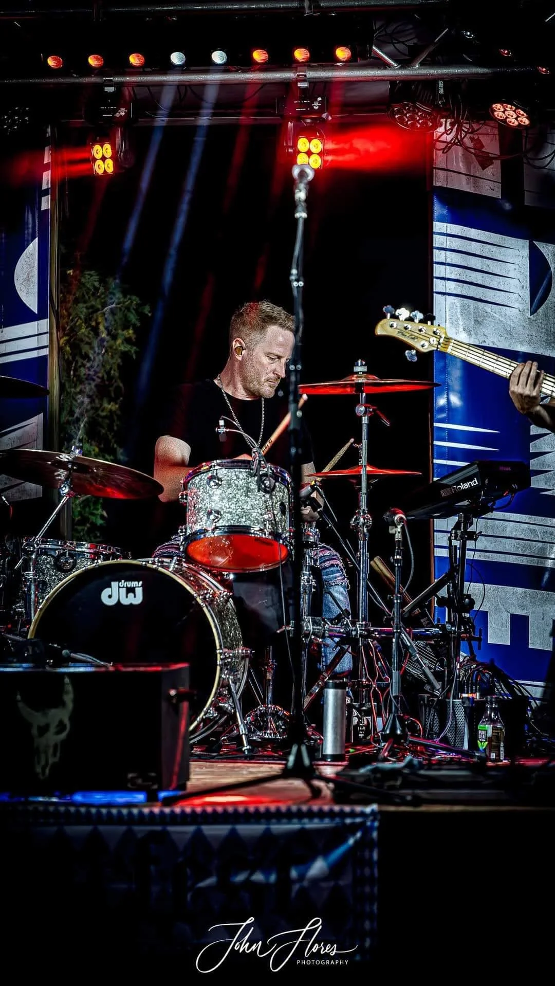 A male drummer playing a drum set on stage, with a bass guitar being played nearby. The stage is lit with red and blue lights, and there is sound equipment and decorations around.