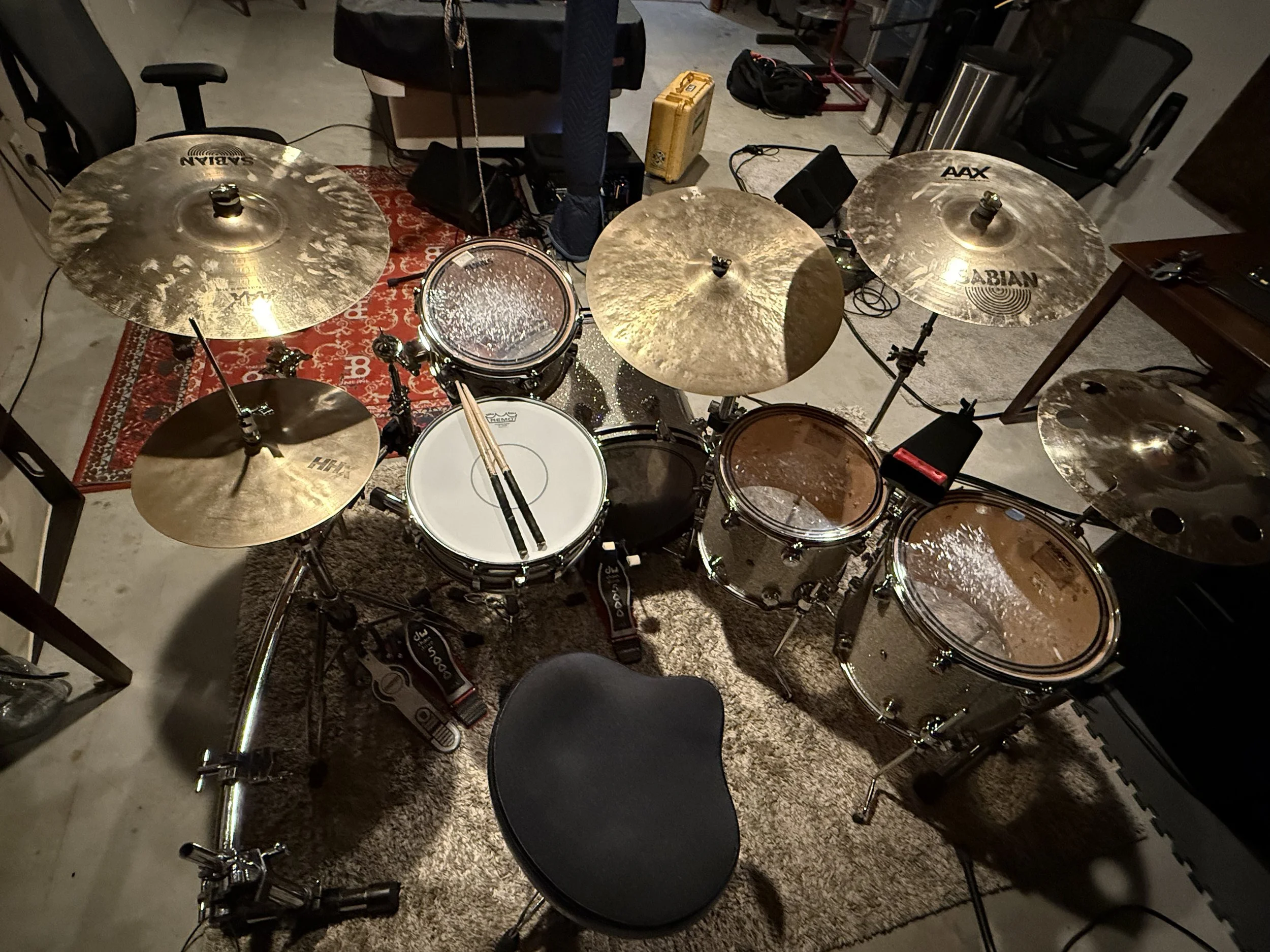 A drum set with multiple cymbals and drums, a stool in front, and various equipment and furniture surrounding it in a room.